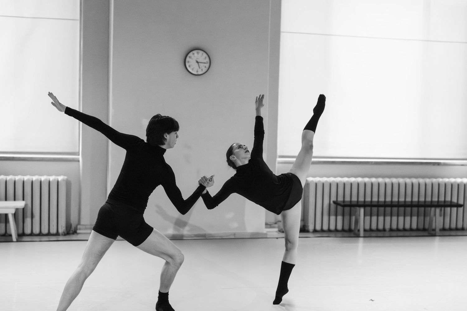 A dancer performs a graceful leap in a bright dance studio with large windows and a ballet barre.