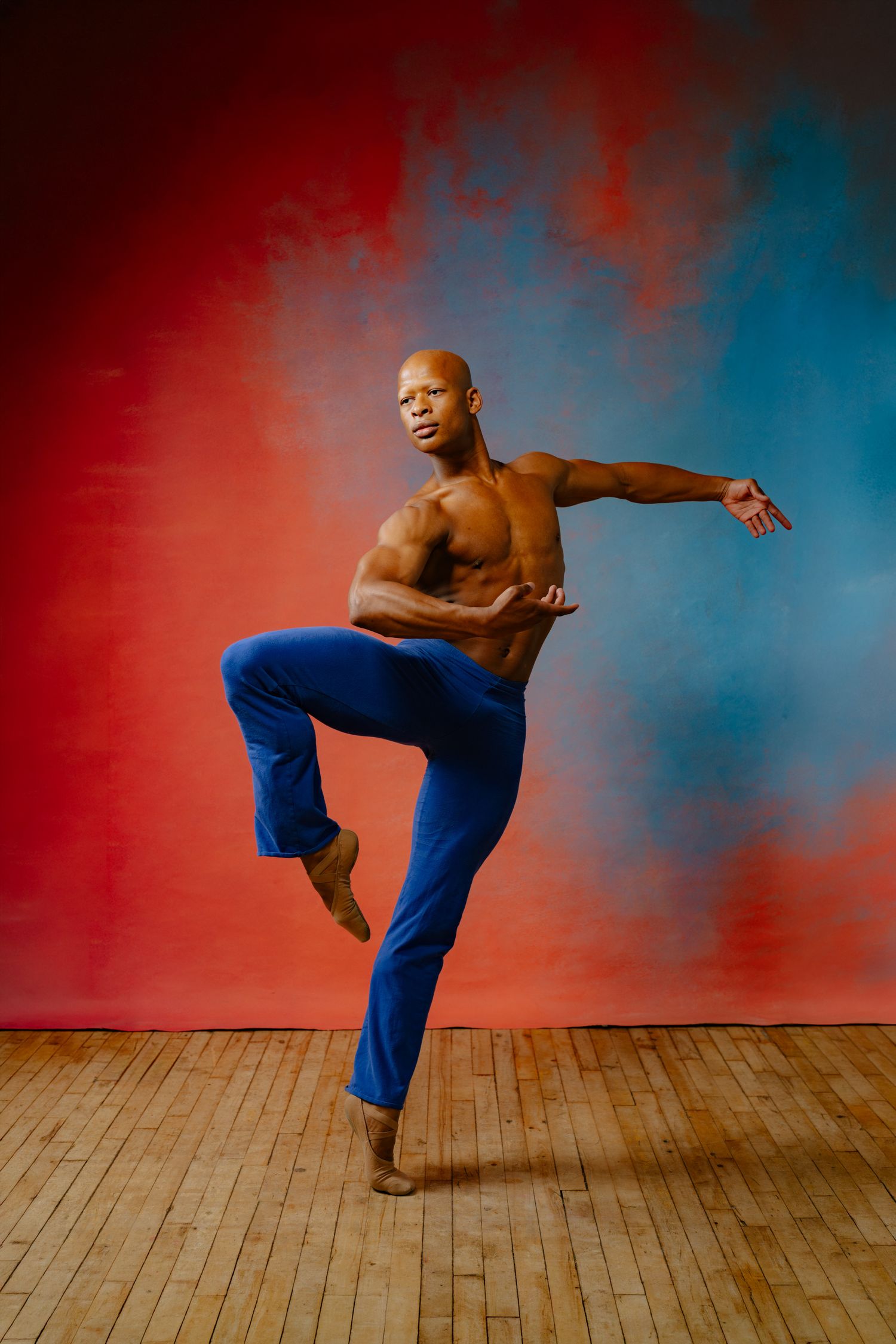 Muscular bald male dancer balancing on one leg, shirtless in blue pants, performing against red and blue backdrop.