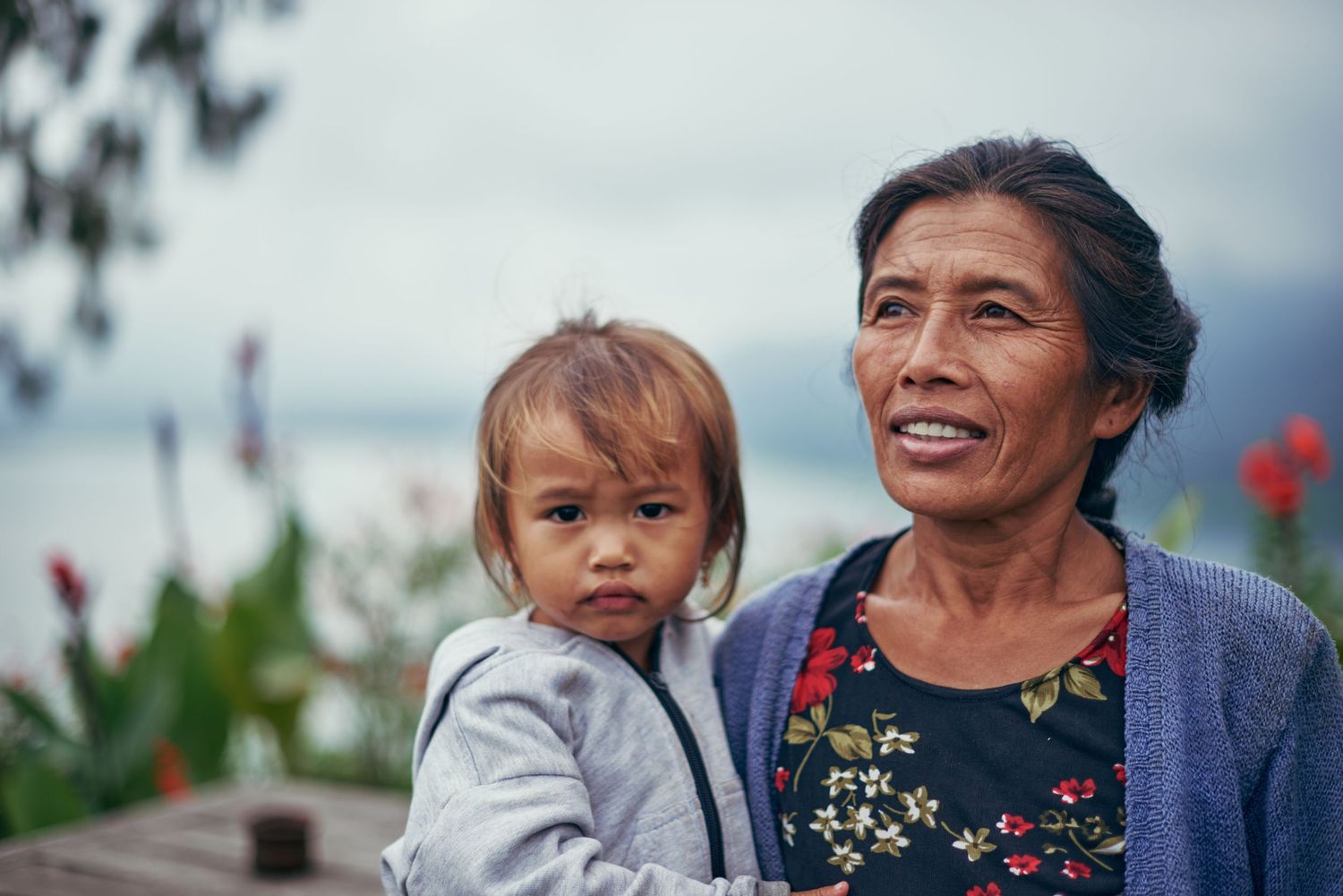 Elderly Asian woman smiling while holding a young toddler outdoors near a lake with flowers in the background.
Lotus Land.Photo