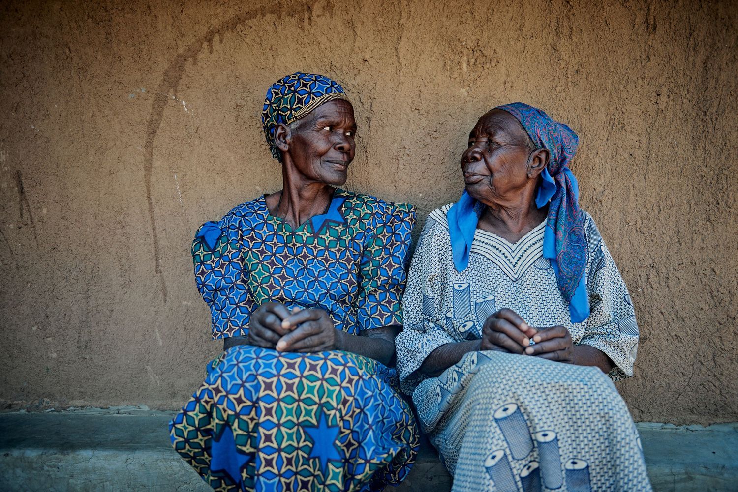 Two elderly African women in colorful traditional attire sit together conversing against a rustic mud wall.