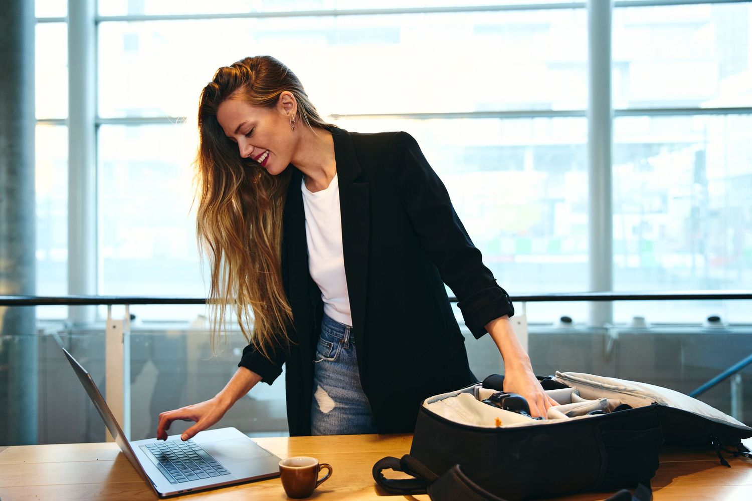 A smiling businesswoman in a black blazer packs her laptop bag while working at a wooden desk near large windows.
Lotus Land.Photo