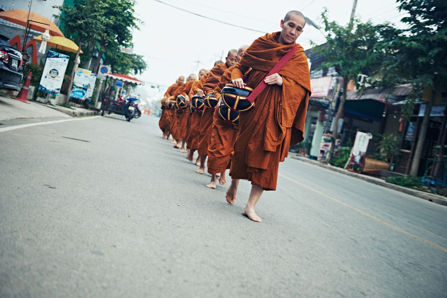 Buddhist monks in orange robes walk barefoot in a line along a street in Southeast Asia.
Lotus Land.Photo