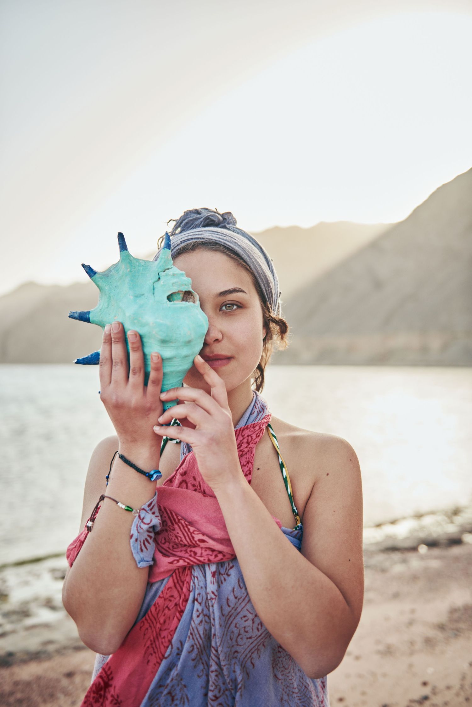 Woman holding a turquoise spiky seashell near her face on a sunny beach with mountains in the background. . Sıdal Ergüder . award-winner