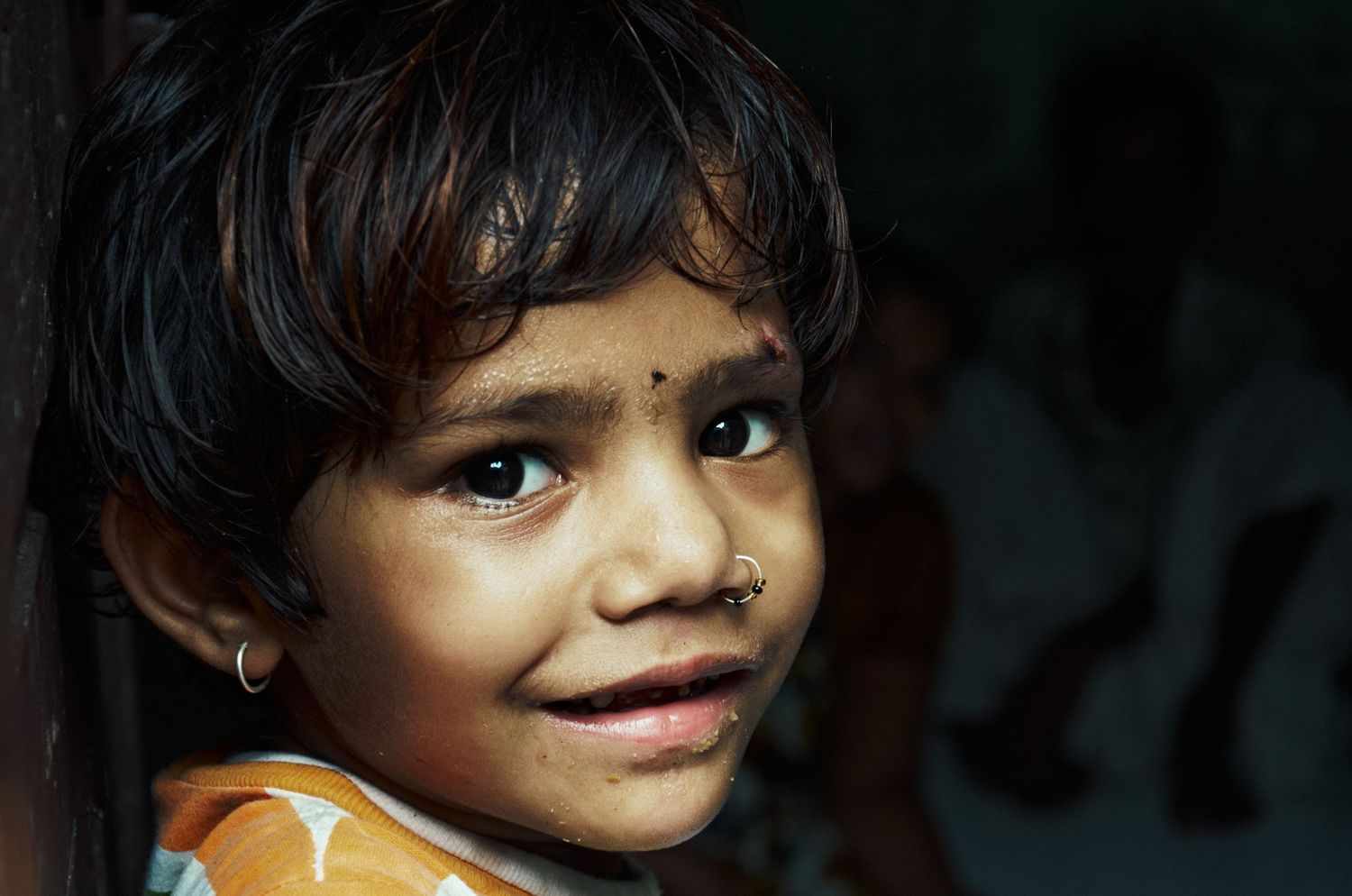 Close-up portrait of a young Indian girl with a nose ring and bindi, smiling shyly at the camera.