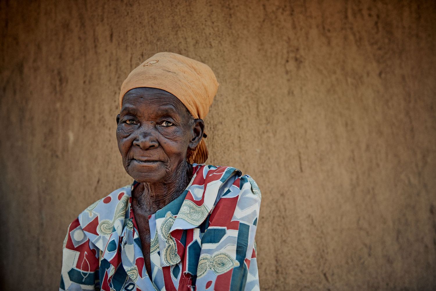 Elderly African woman wearing colorful patterned shirt and orange headwrap standing against a mud wall.