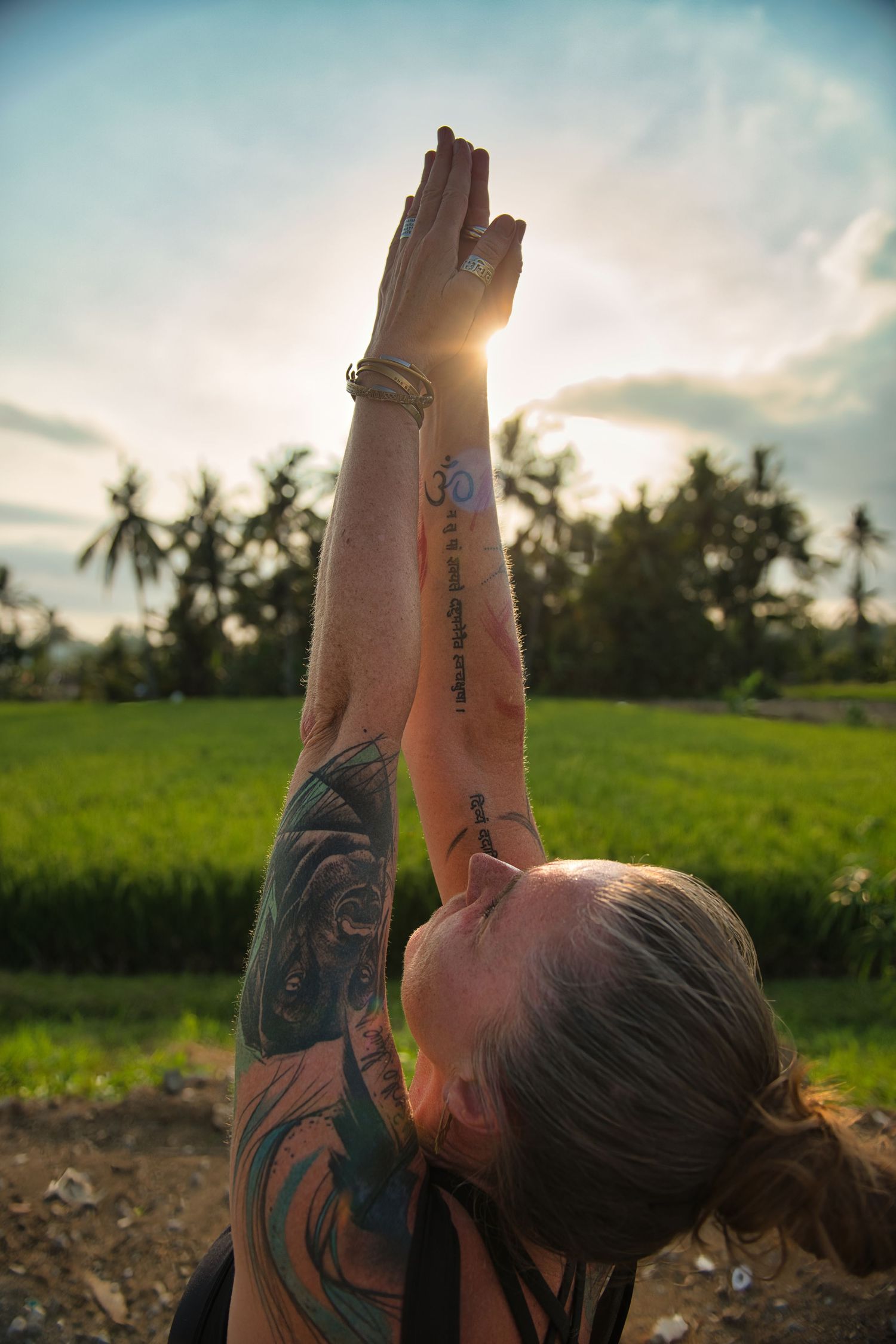 Tattooed woman practicing yoga with hands raised in prayer pose against a lush green tropical landscape at sunset.
Lotus Land.Photo