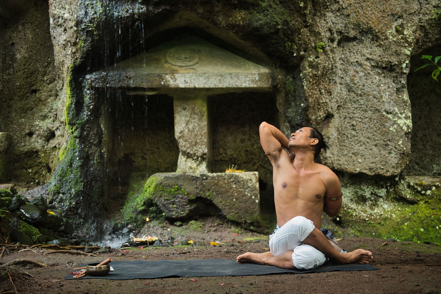 Shirtless man practicing yoga in a serene outdoor setting near mossy stone ruins and a waterfall.
Lotus Land.Photo