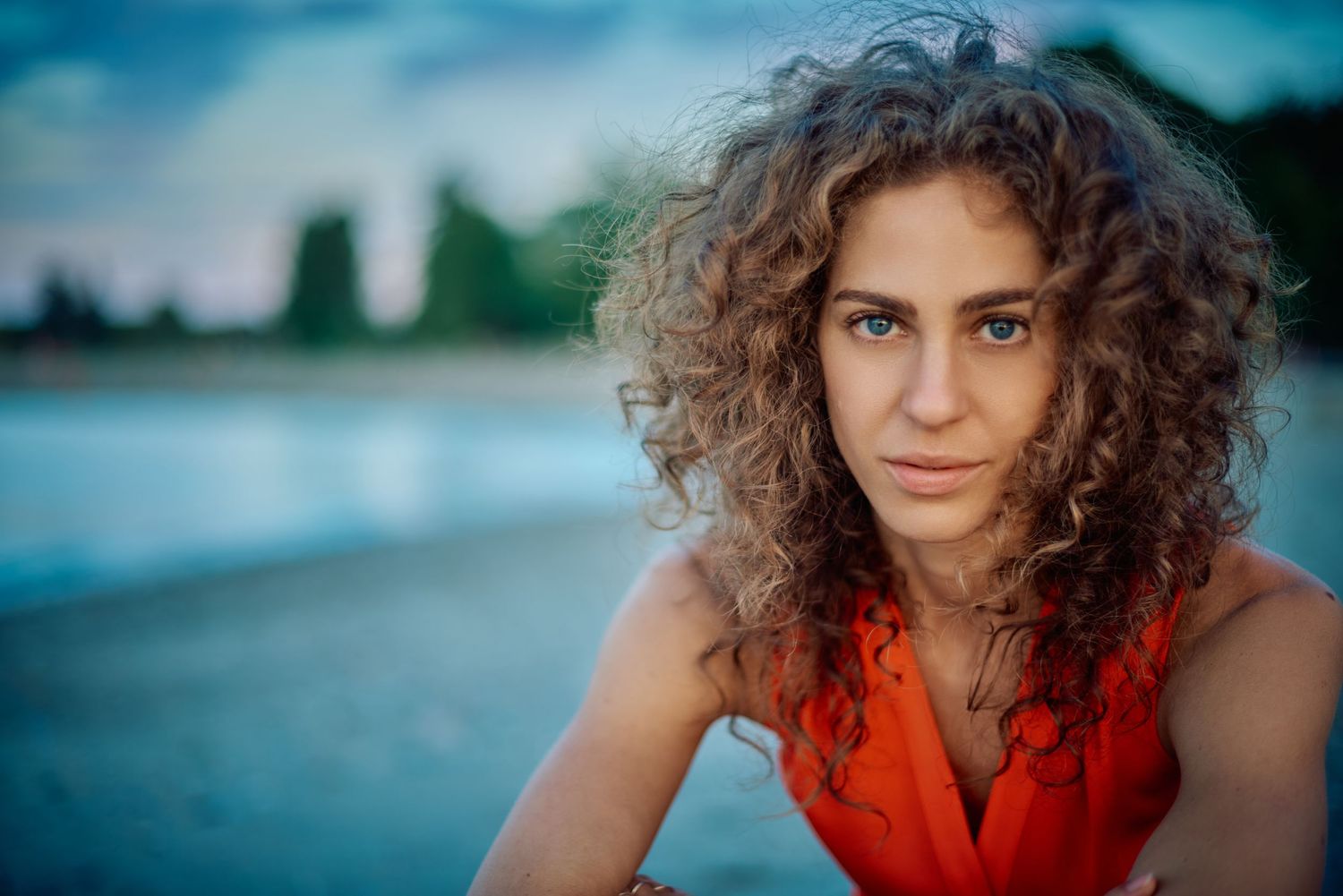 Young woman with curly hair wearing an orange top poses outdoors near water at dusk.
Carmel Amit
Lotus Land.Photo