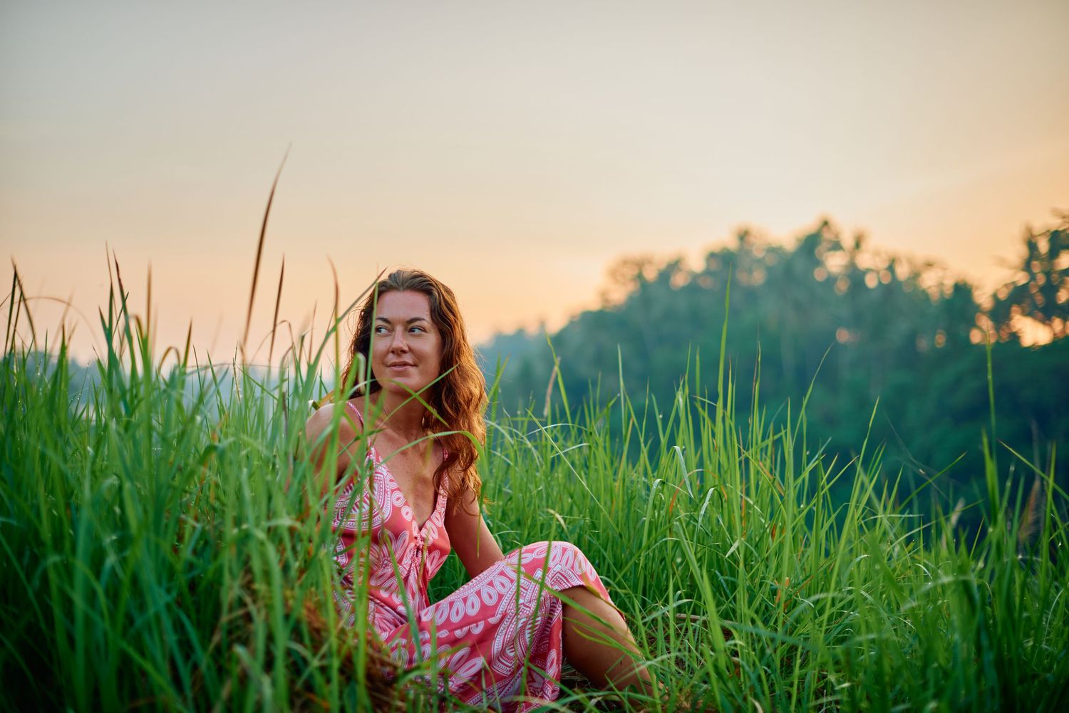 Woman in pink floral dress sitting in tall green grass during golden hour sunset with trees in background.
Lotus Land.Photo