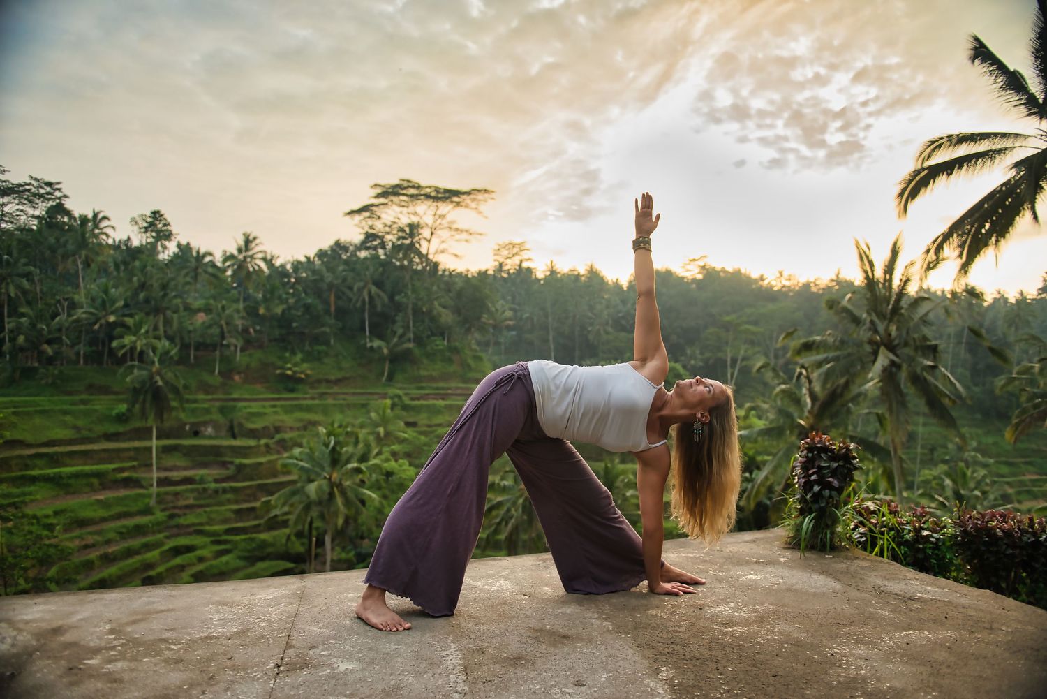 Woman practicing yoga in a triangle pose on a stone terrace surrounded by lush green rice terraces at sunset.
Lotus Land.Photo