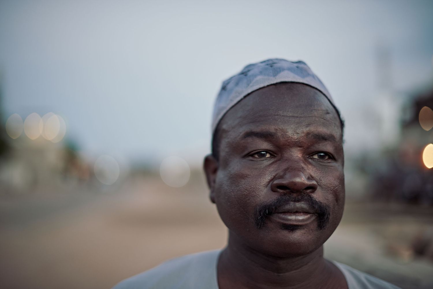 Portrait of a middle-aged African man wearing a traditional kufi cap at dusk on a blurred street background.