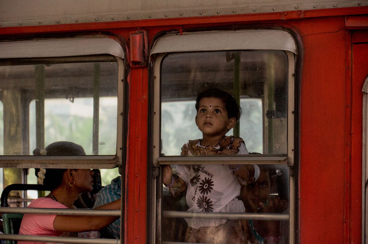 Young child gazing out of a red train window, with passengers visible inside the colorful vintage railcar.