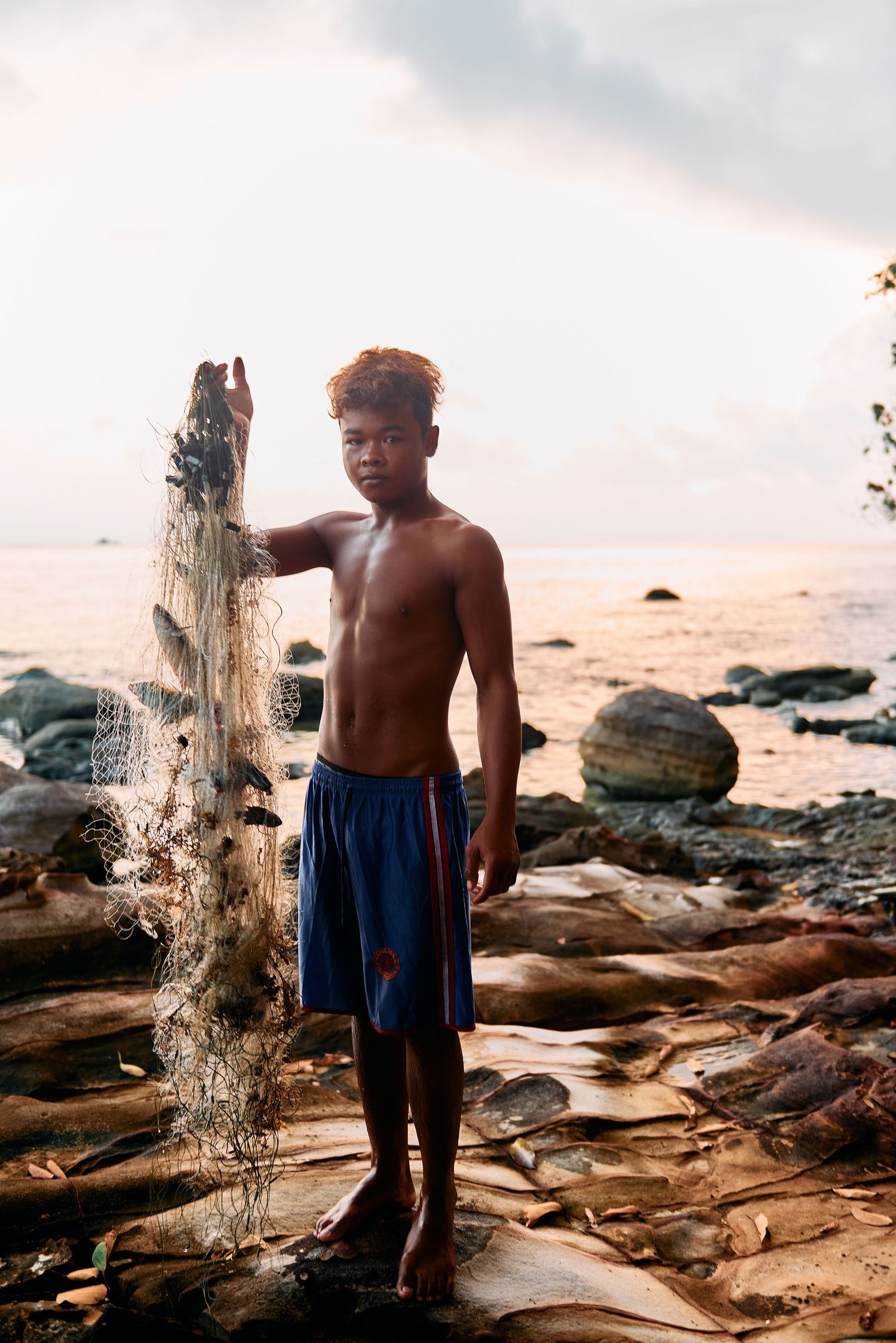 A shirtless boy stands on rocky shoreline holding a fishing net at sunset.
Lotus Land.Photo
