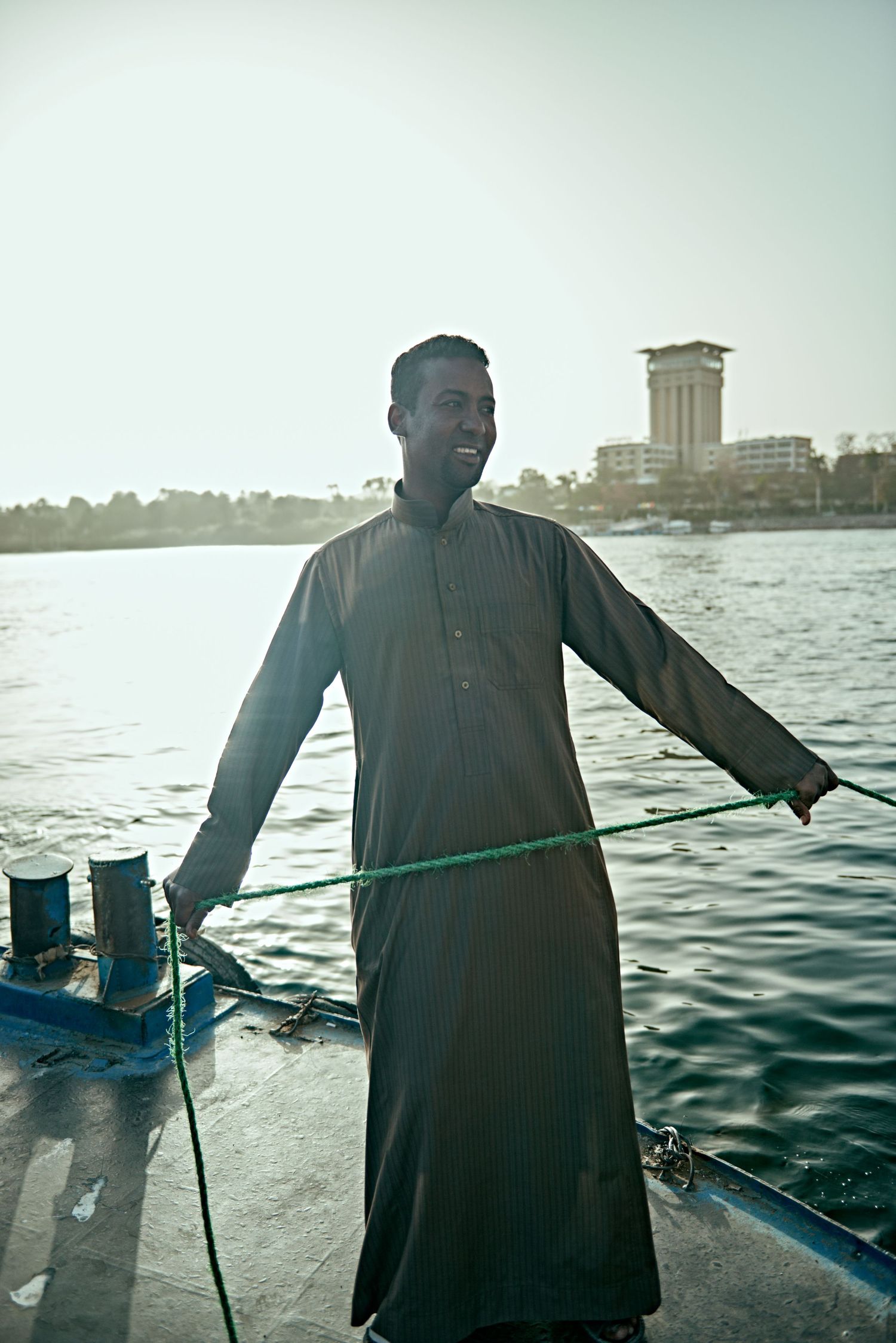 Smiling man in traditional dark robe holding rope on a boat with river and tower in background.