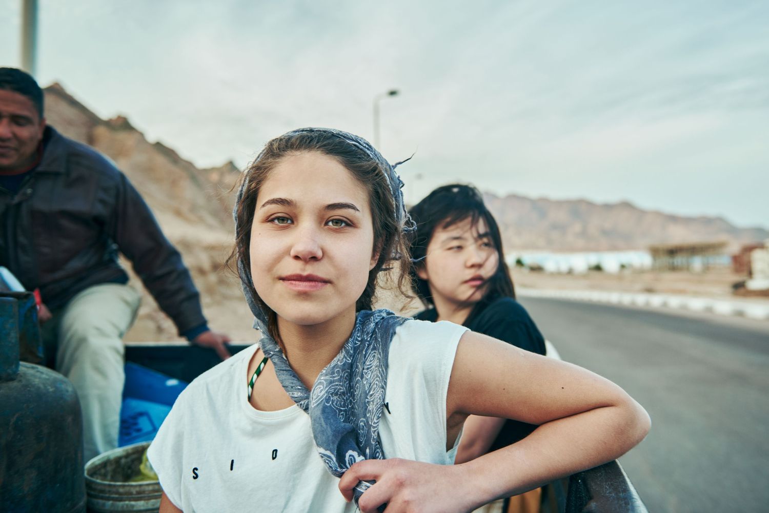 Two young women sitting on a truck bed on a desert road with mountains in the background. Sıdal Ergüder . multi-award-winning director