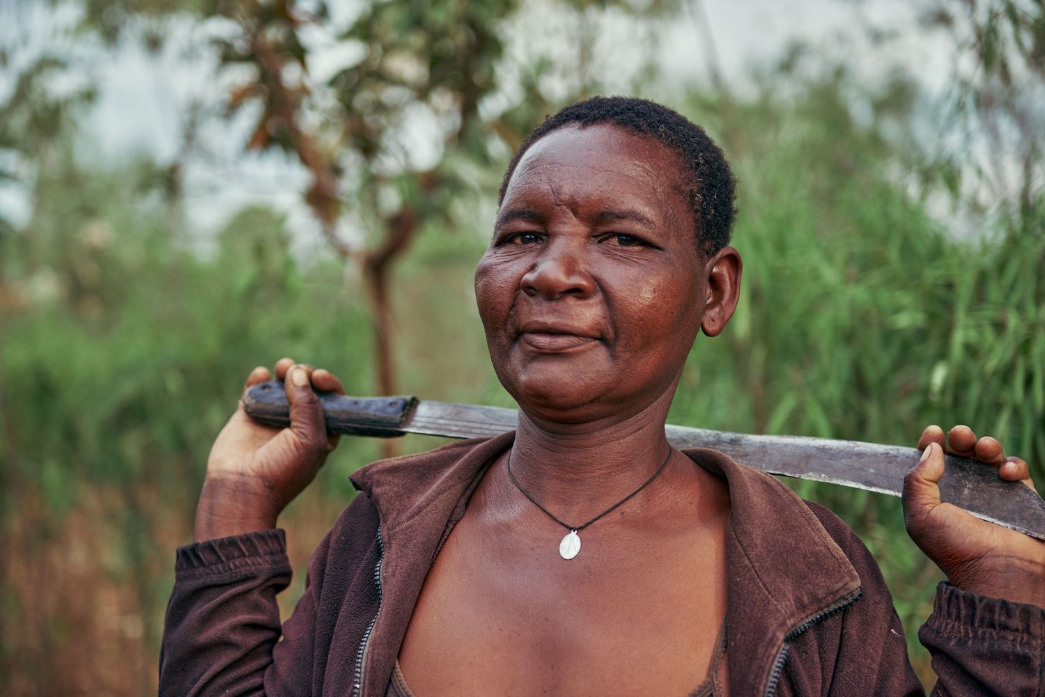 African woman balancing a tool on her shoulders outdoors, with lush green trees in the background.