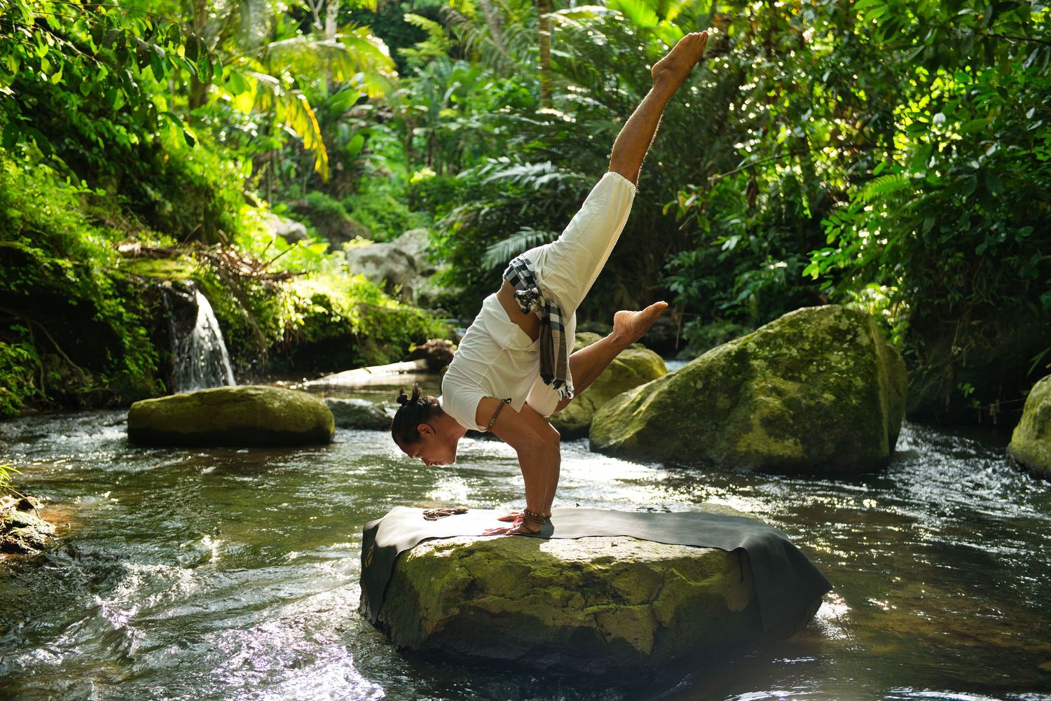 Person performing a handstand yoga pose on a rock in a lush tropical stream surrounded by green moss and waterfalls.
Lotus Land.Photo