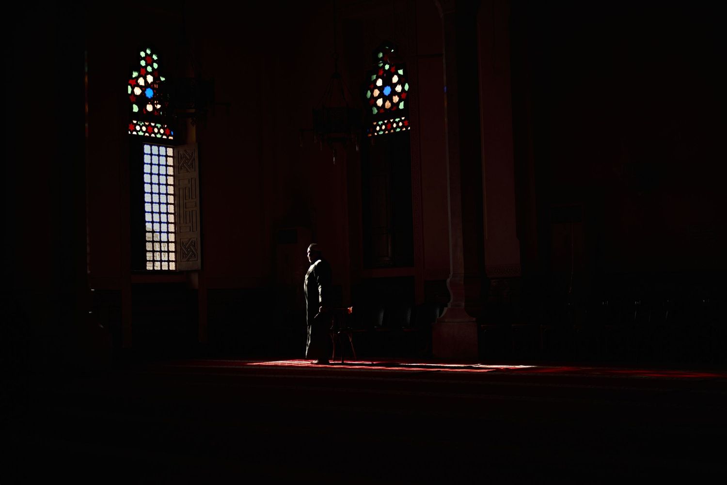 A solitary figure stands in a dimly lit mosque, illuminated by colorful stained glass windows casting light on the floor.