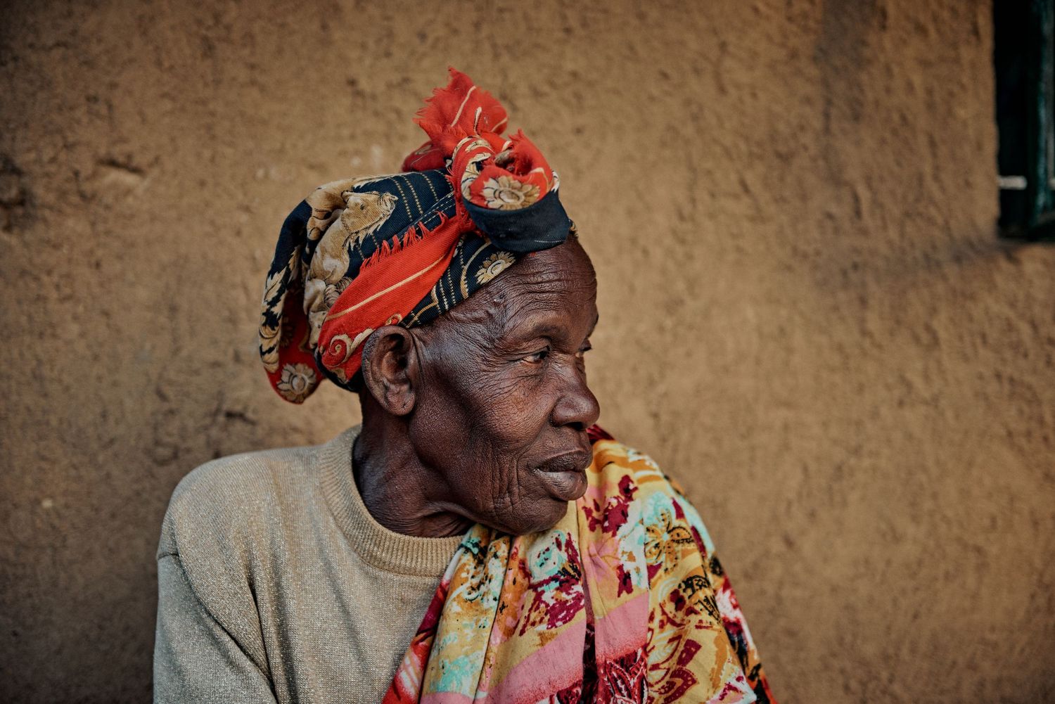 Elderly African woman wearing colorful floral scarf and red headwrap, seated outdoors near a rustic mud wall.