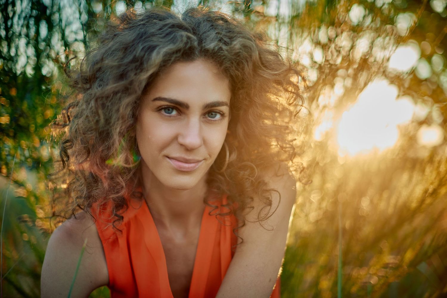 Young woman with curly hair in orange top smiling softly outdoors in golden sunlight among green foliage.
Carmel Amit
Lotus Land.Photo