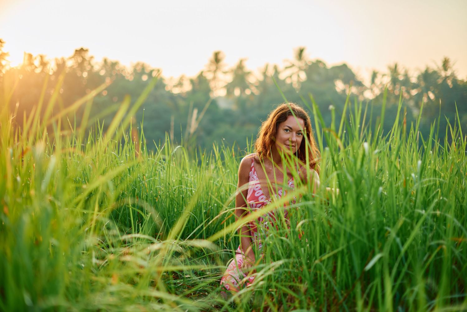 A young woman in a floral dress sits among tall green rice fields at golden sunset with tropical palms in background.
Lotus Land.Photo