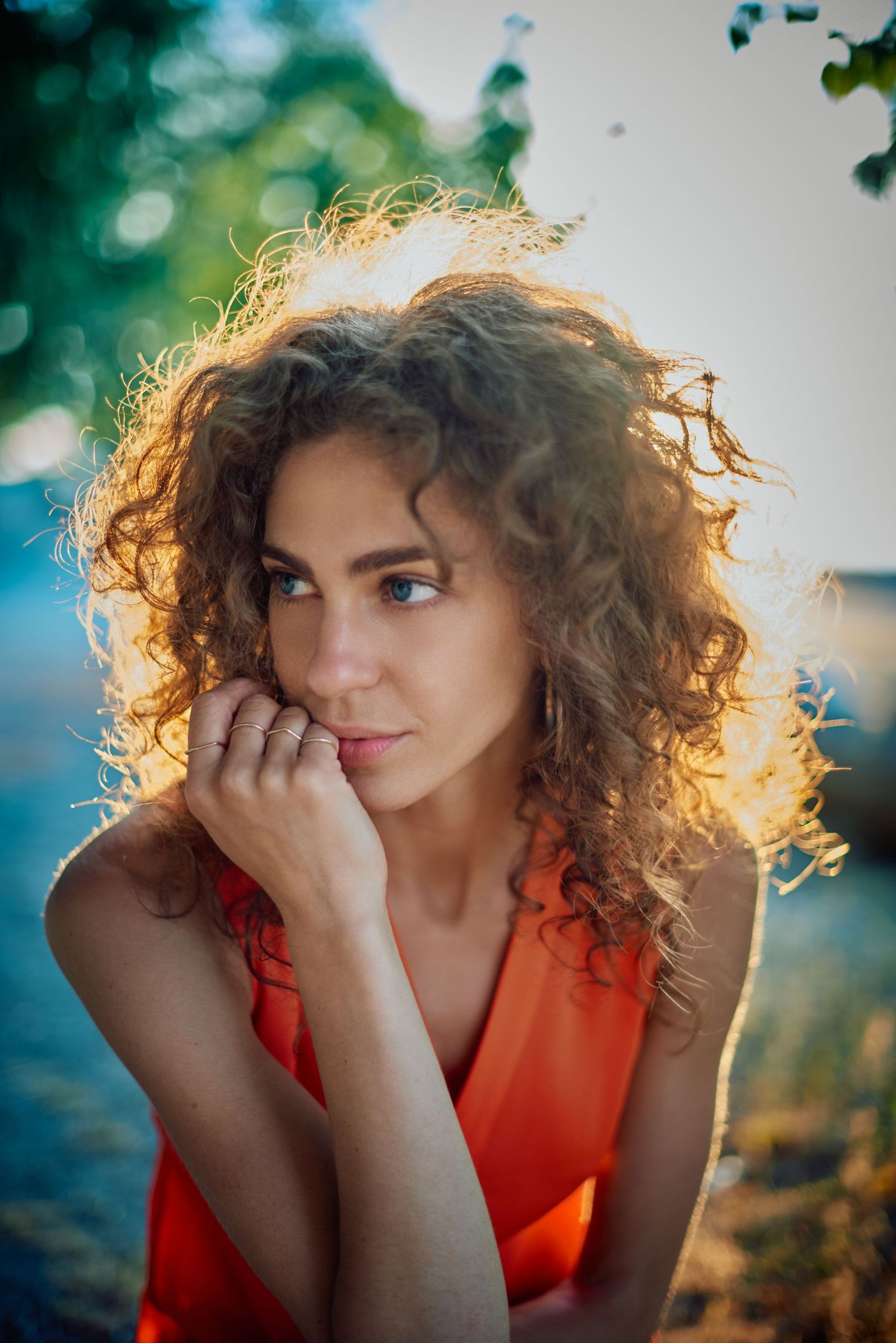A young woman with curly auburn hair in an orange dress gazes, bathed in warm golden sunlight outdoors.
Carmel Amit
Lotus Land.Photo