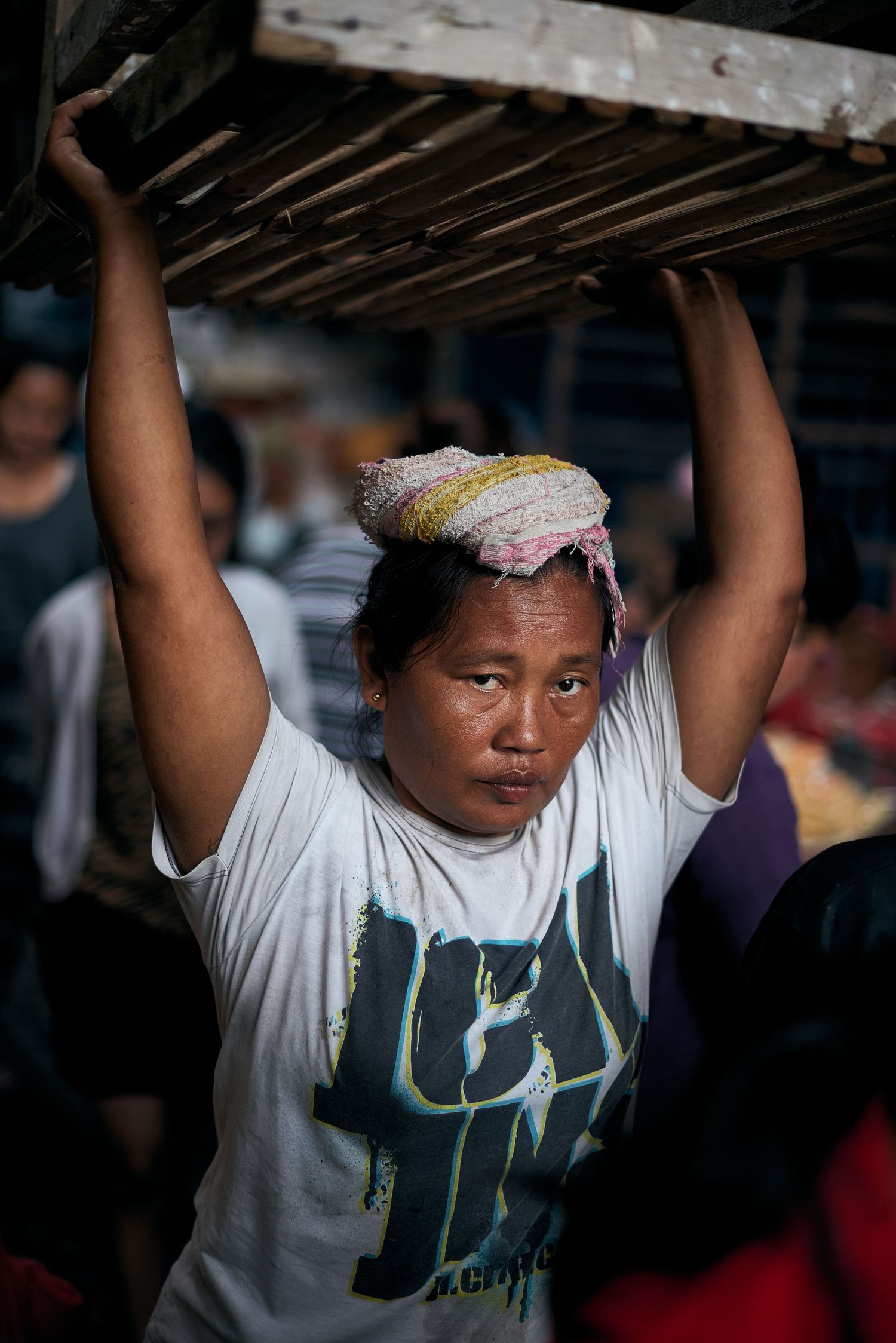 Woman in white t-shirt carrying wooden crate on her head at a busy market.
Lotus Land.Photo