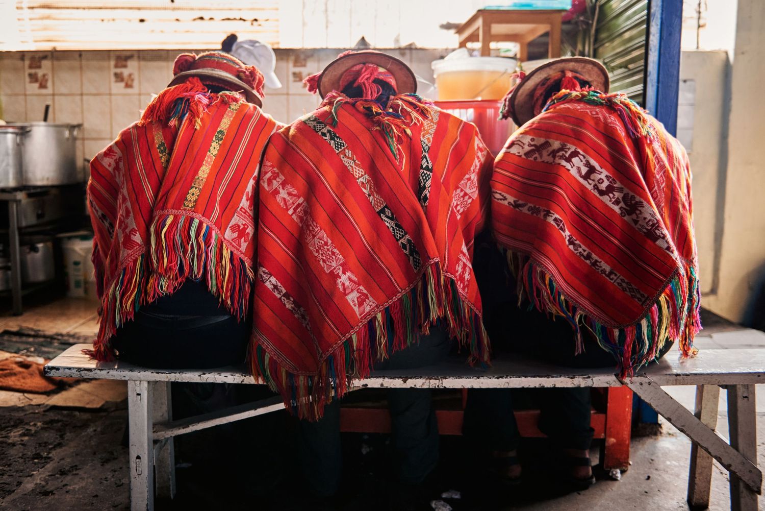 Three people wearing traditional red ponchos and hats sit at a table in a rustic kitchen setting.
Lotus Land.Photo