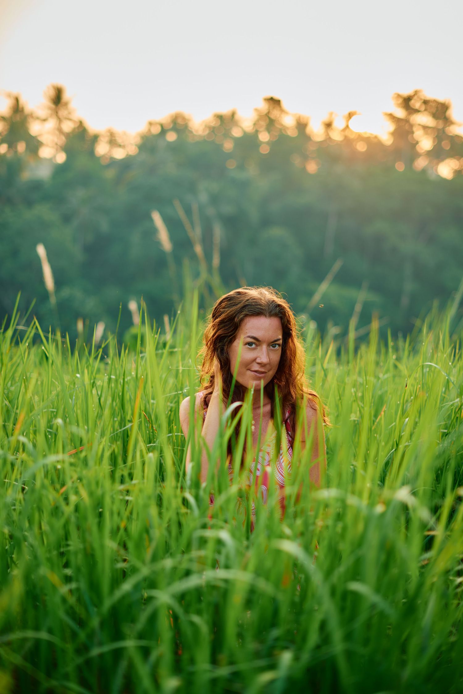 Woman with auburn hair sitting in lush green rice field at golden sunset with trees in background.
Lotus Land.Photo