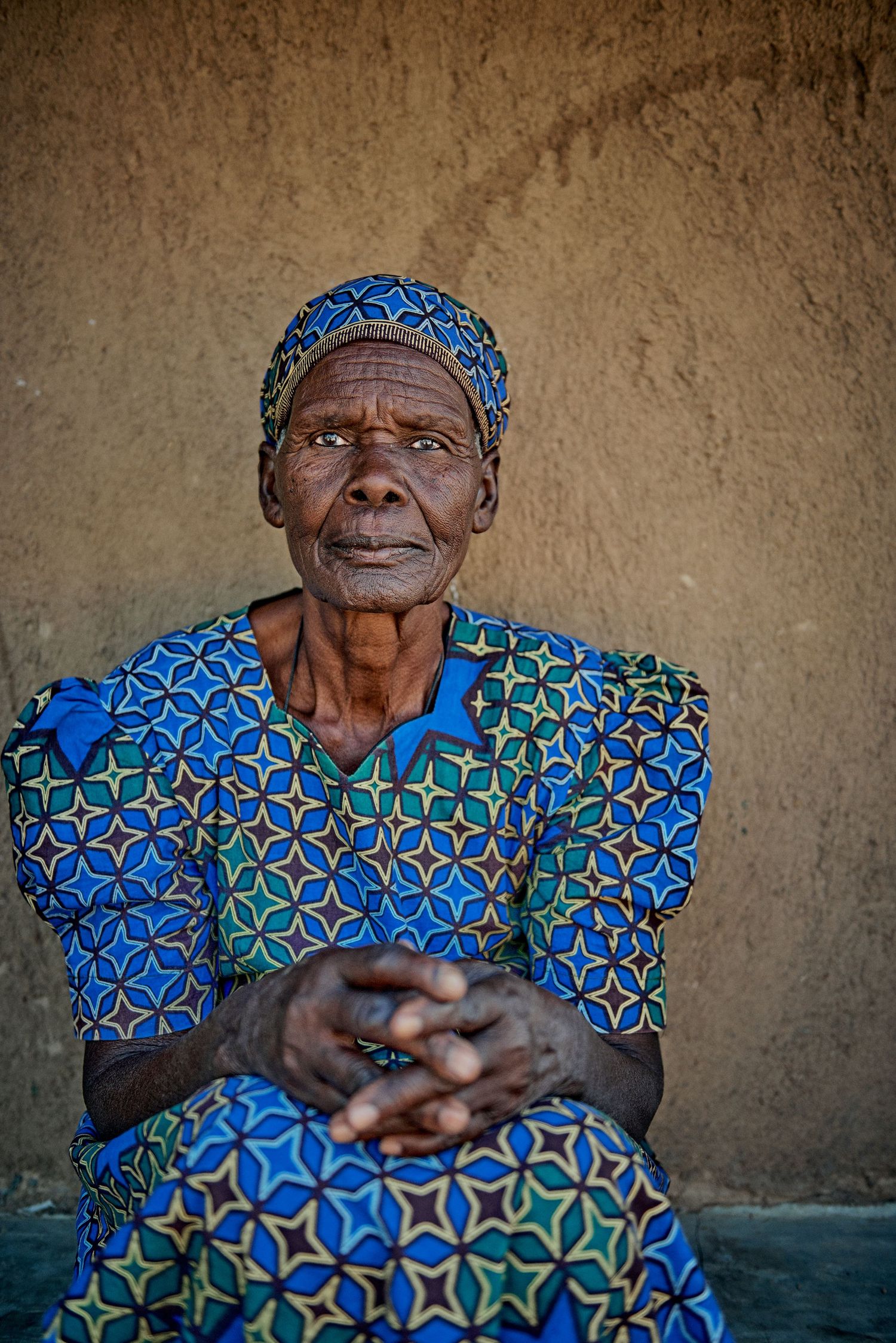 Elderly African woman in vibrant blue patterned dress and headwrap sits with clasped hands against a mud wall.