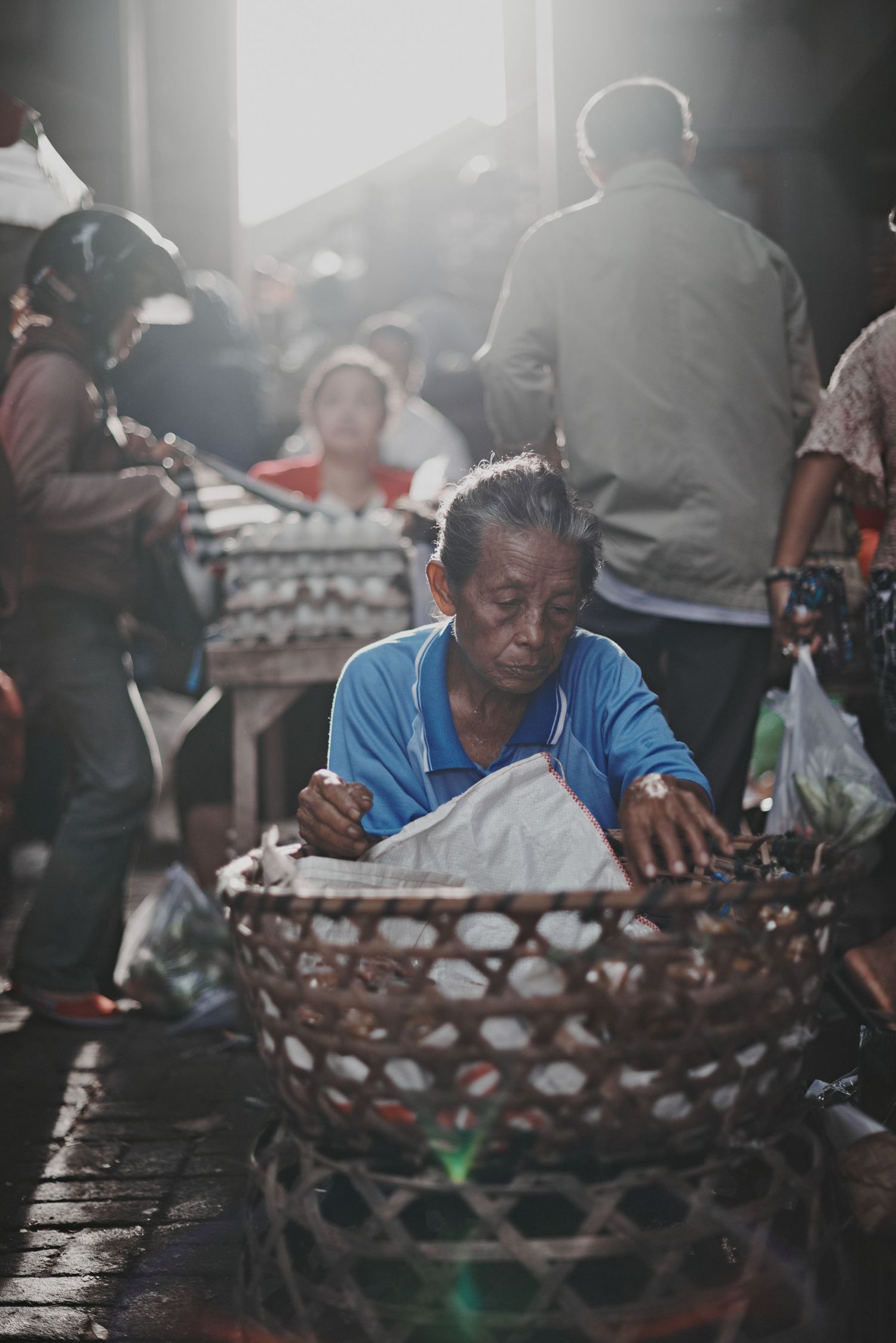 Elderly man in blue shirt sorting goods in woven basket at busy Asian outdoor market with soft backlit sunlight.
Lotus Land.Photo