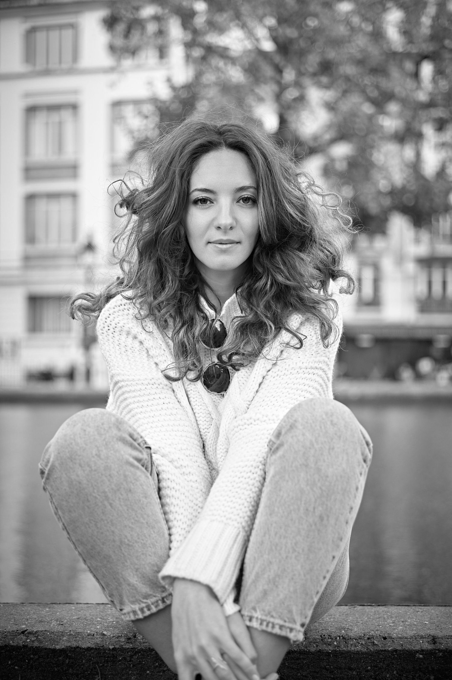 Black and white portrait of a young woman with curly hair sitting cross-legged outdoors at waterfront.
Kenza Kassabi Tedx
Lotus Land.Photo
