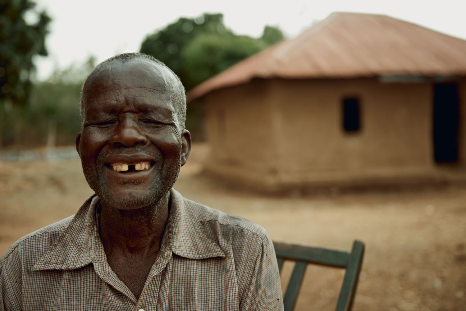 Smiling elderly African man with missing teeth sits outdoors near a traditional round mud hut with thatched roof.