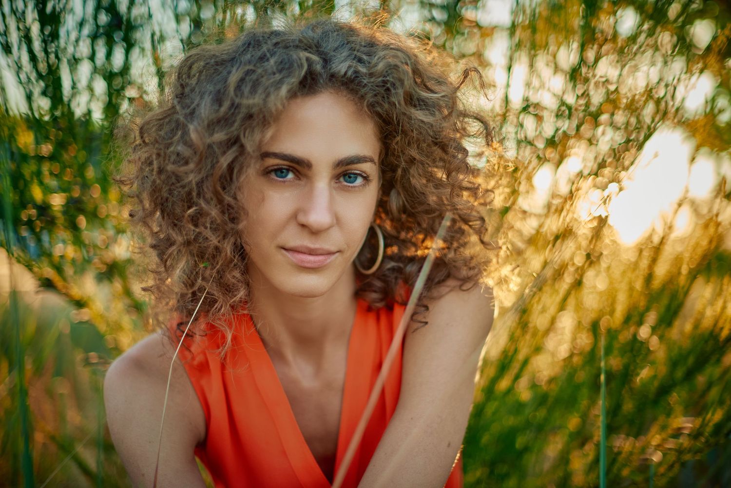 Woman with curly hair in orange top poses outdoors among tall green grass in golden sunset light.
Carmel Amit
Actress
Lotus Land.Photo