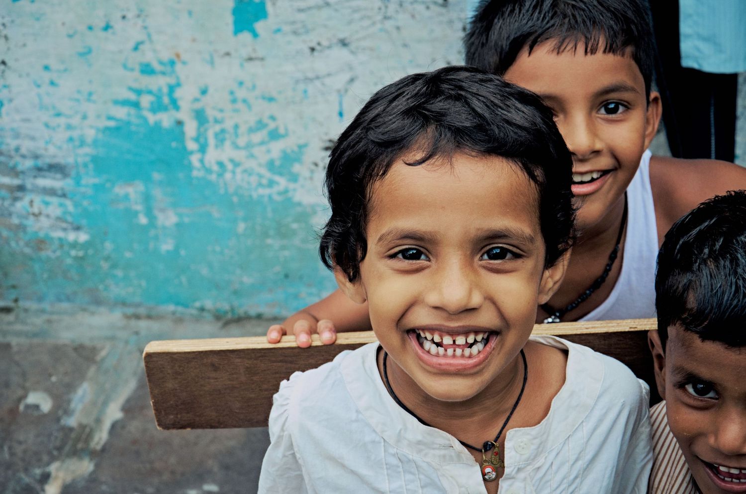 Smiling Indian children playing outdoors near a turquoise wall, with a boy in front holding a wooden stick.