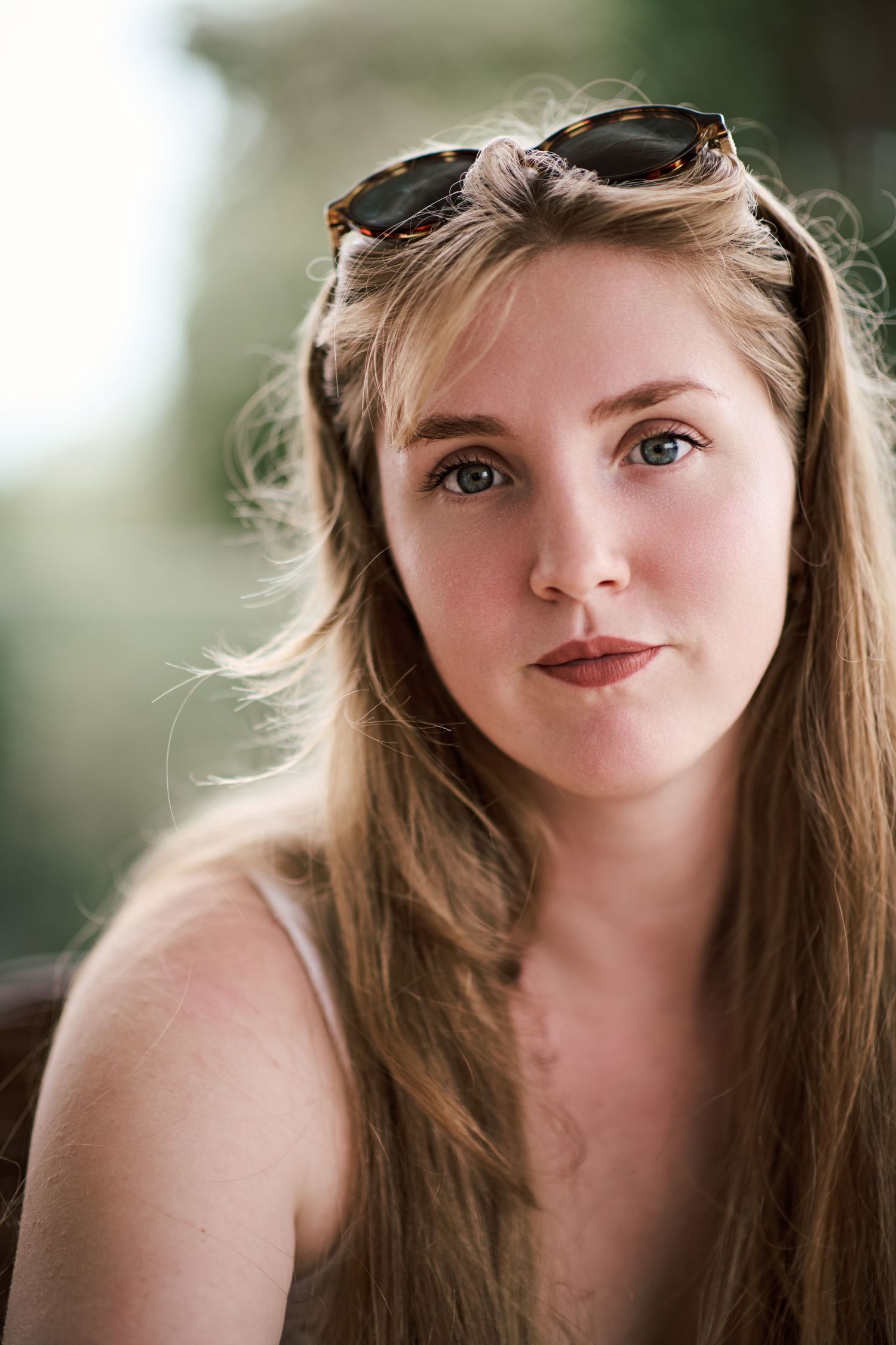 Young woman with long blonde hair and sunglasses on her head gazes thoughtfully outdoors in natural light.
