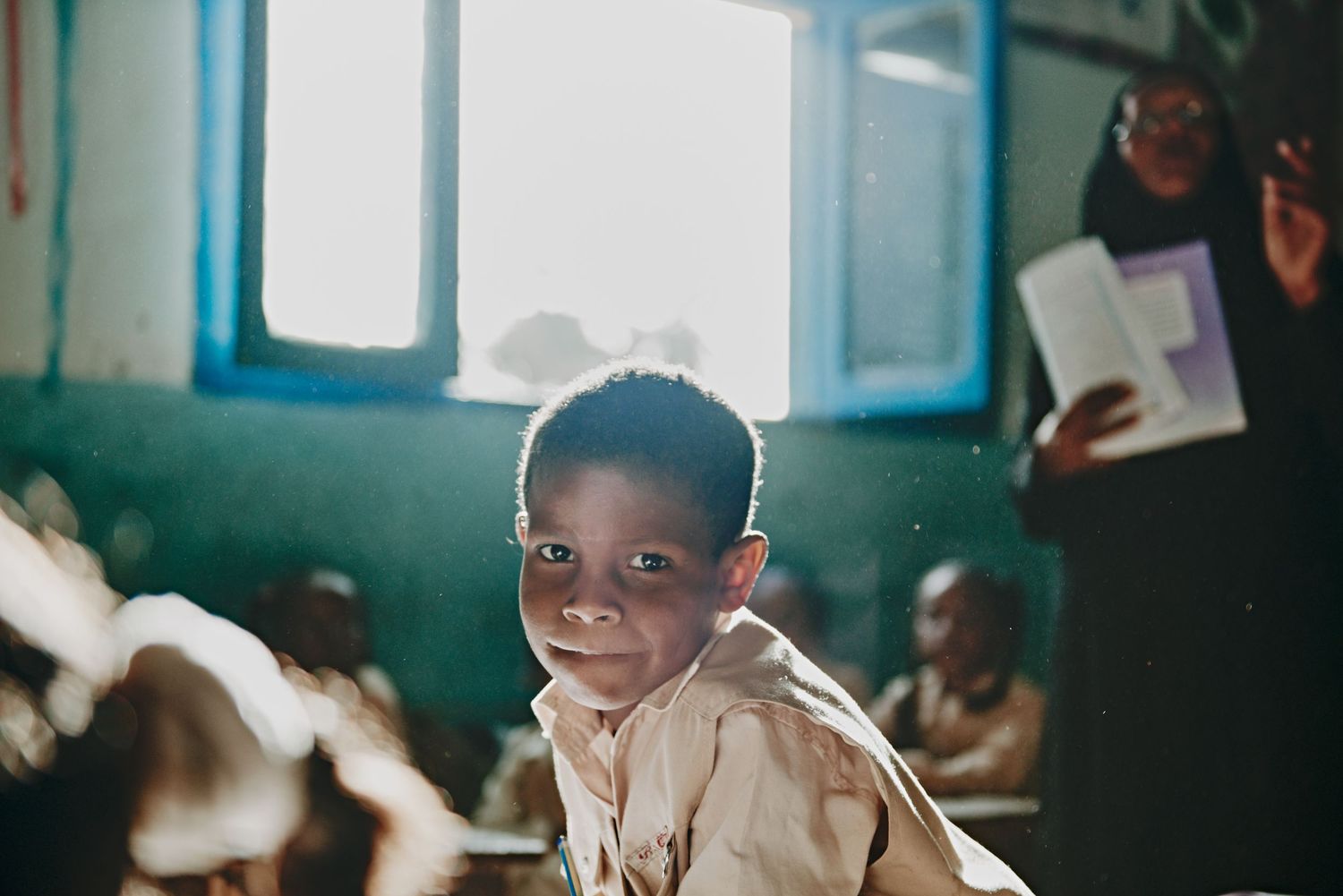 Young African boy smiling at camera in a classroom setting with other children and a teacher in background.