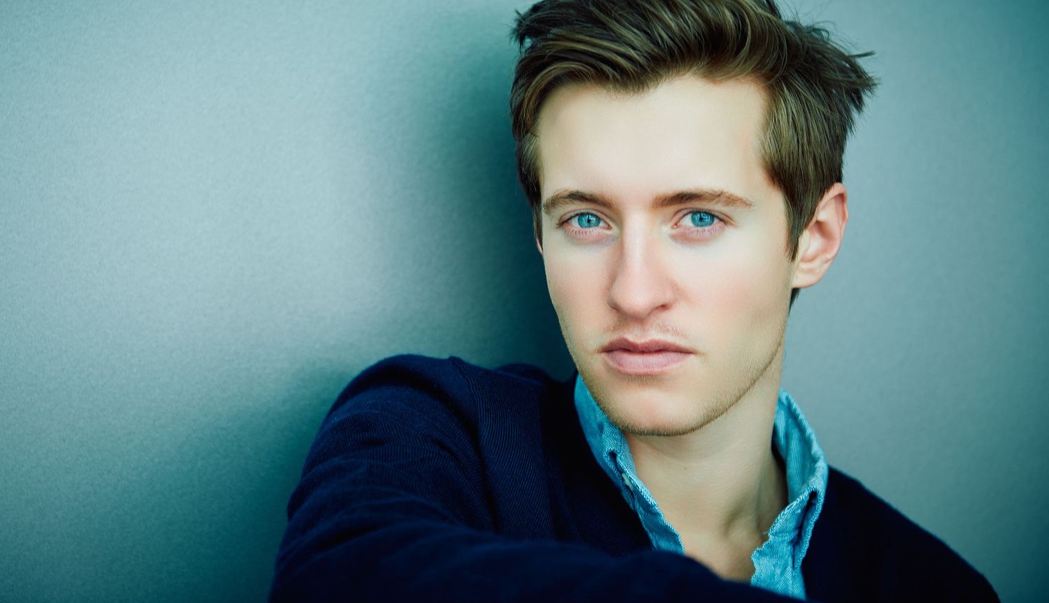 Young man with blue eyes and short brown hair posing against a gray wall in a navy blazer.
Lotus Land.Photo