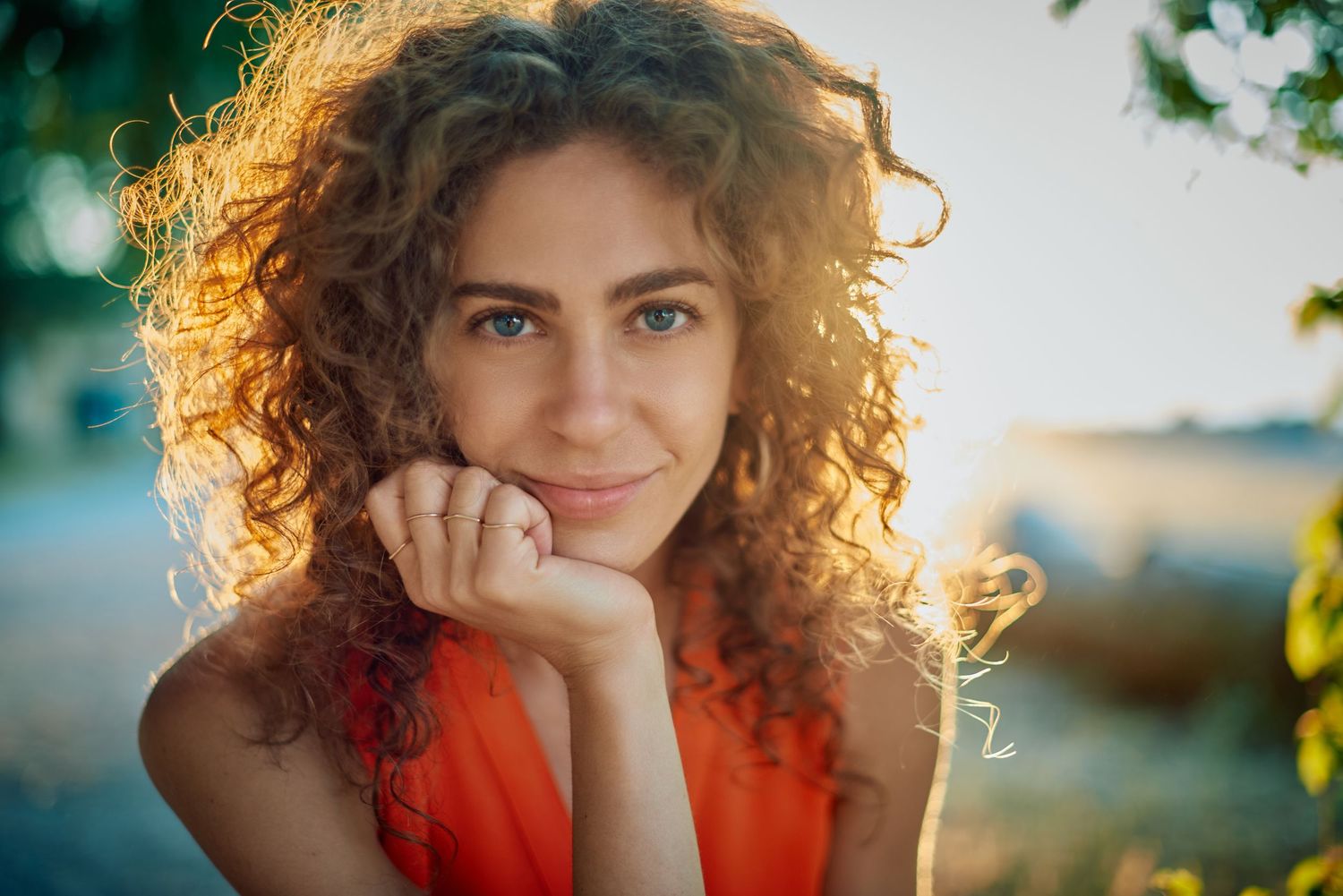 Young woman with curly auburn hair and blue eyes smiling outdoors warm golden sunlight wearing an orange top.
Carmel Amit
Lotus Land.Photo