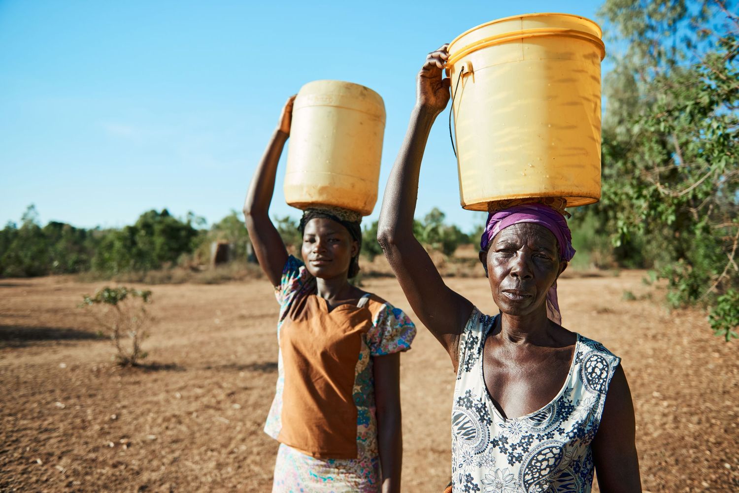 Two African women carrying large yellow water buckets on their heads across dry, arid land.