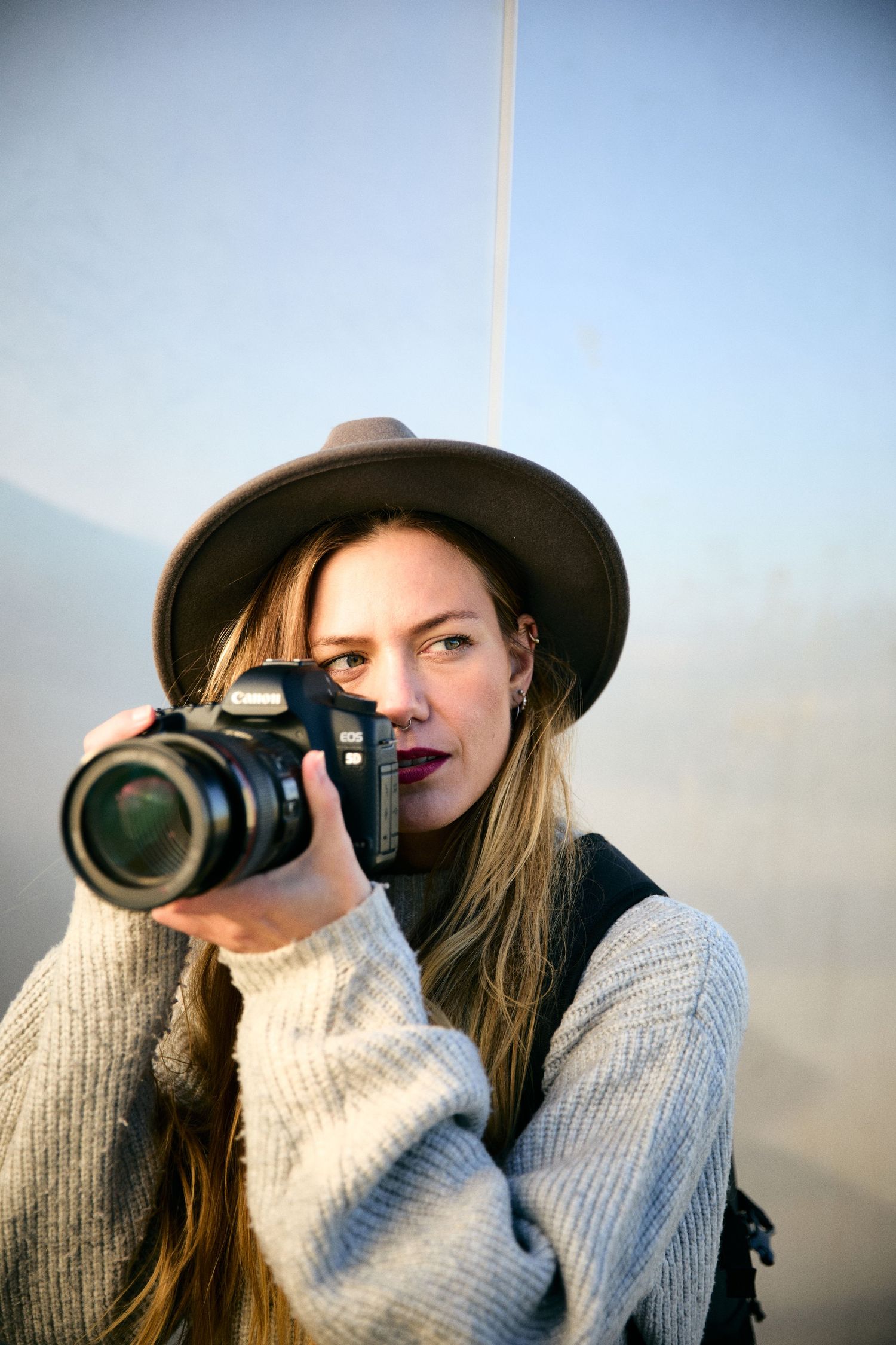 Woman in brown hat holding DSLR camera up to her face outdoors in natural light wearing a cozy sweater.
Lotus Land.Photo