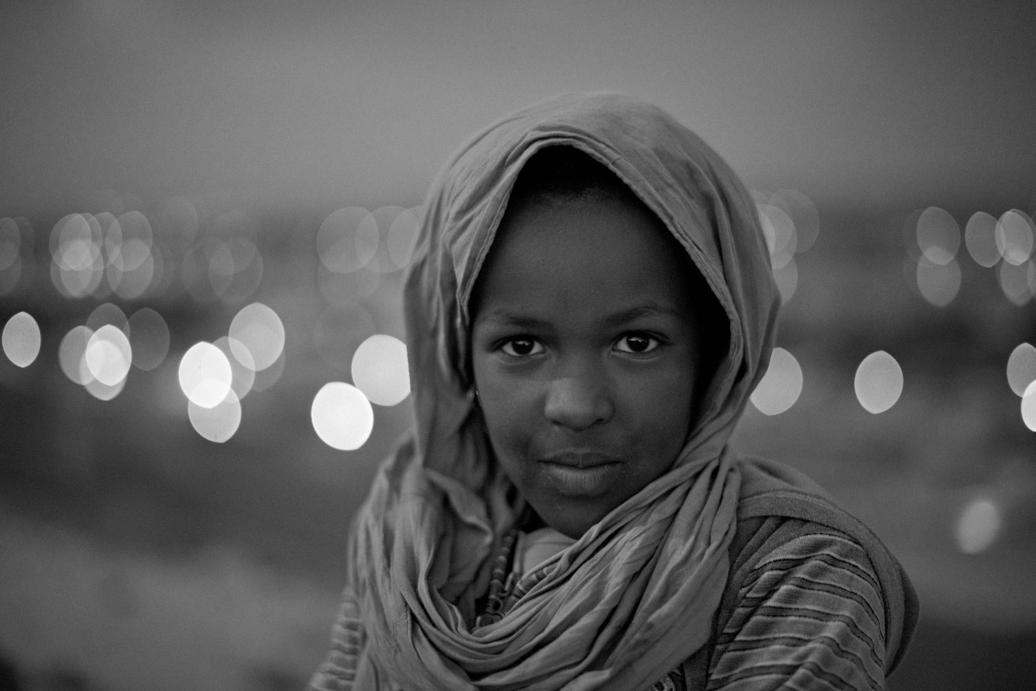 Black and white portrait of a young woman wearing a hijab with bokeh city lights in the background.