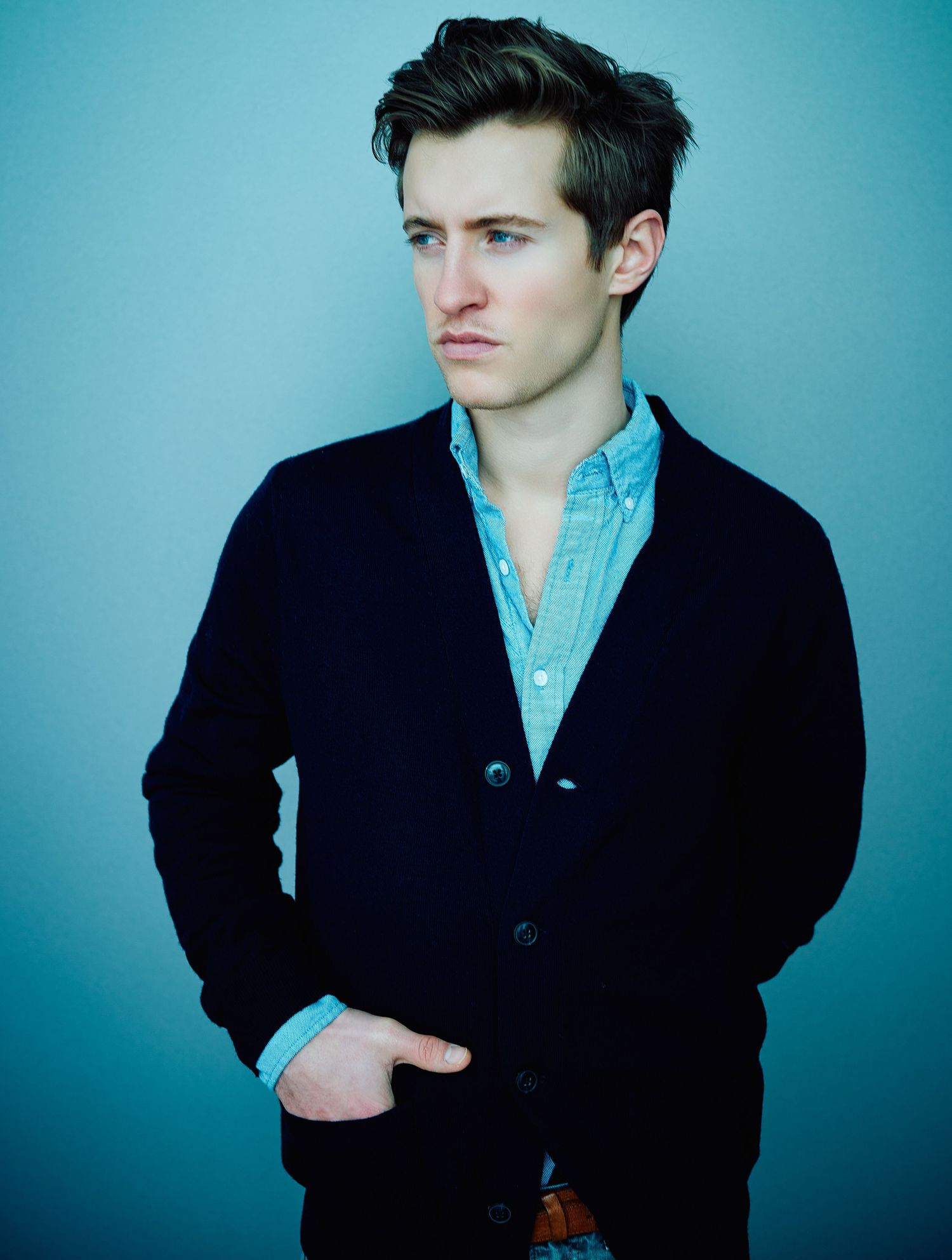 Young man in navy cardigan and light blue shirt posing against a teal wall with hands in pockets.
Lotus Land.Photo