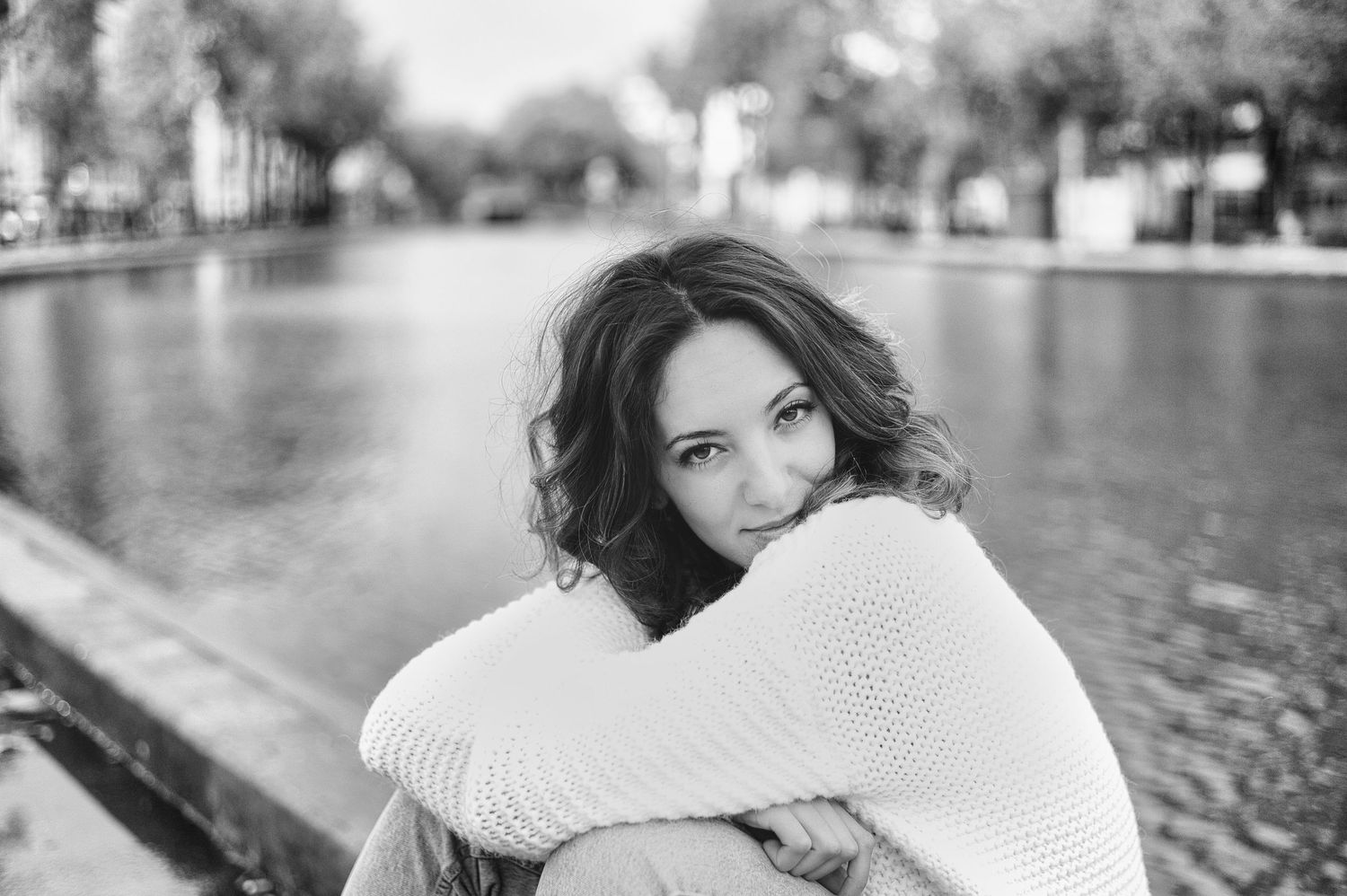 Black and white portrait of a smiling woman in a knit sweater sitting by a canal, hugging her knees.
Kenza Kassabi Tedx
Lotus Land.Photo