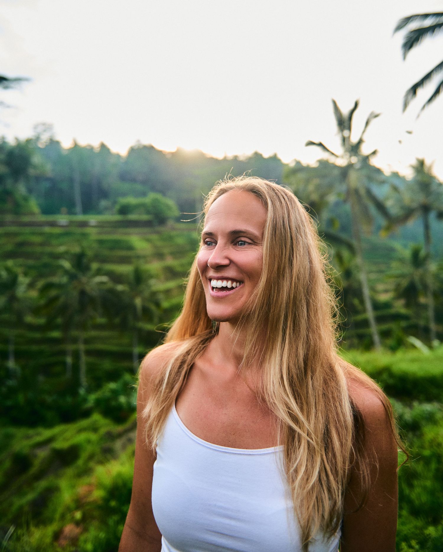 Smiling blonde woman in white tank top at lush green Bali rice terraces with palm trees at sunset.
Lotus Land.Photo