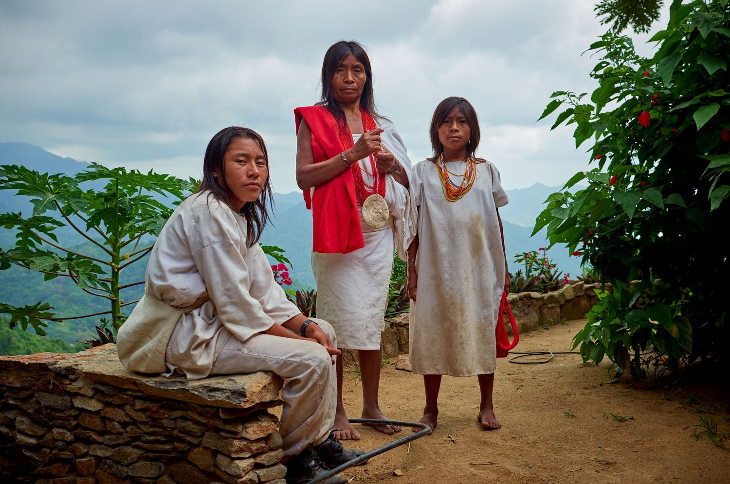 Three indigenous women in traditional white and red attire stand outdoors on a hillside with lush green plants.
Lotus Land.Photo