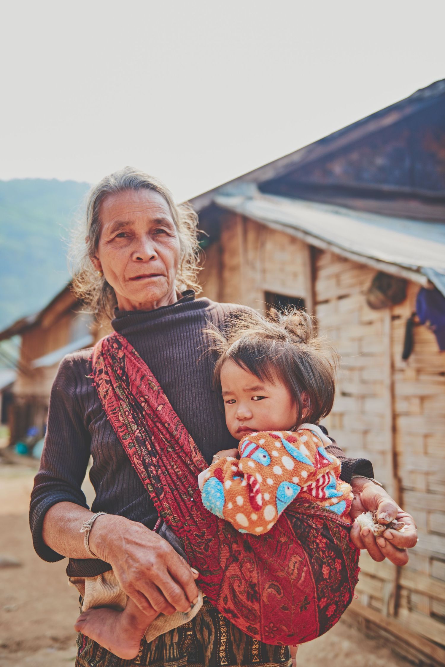 Elderly woman carrying a young child in a colorful sling in a rural Southeast Asian village setting.