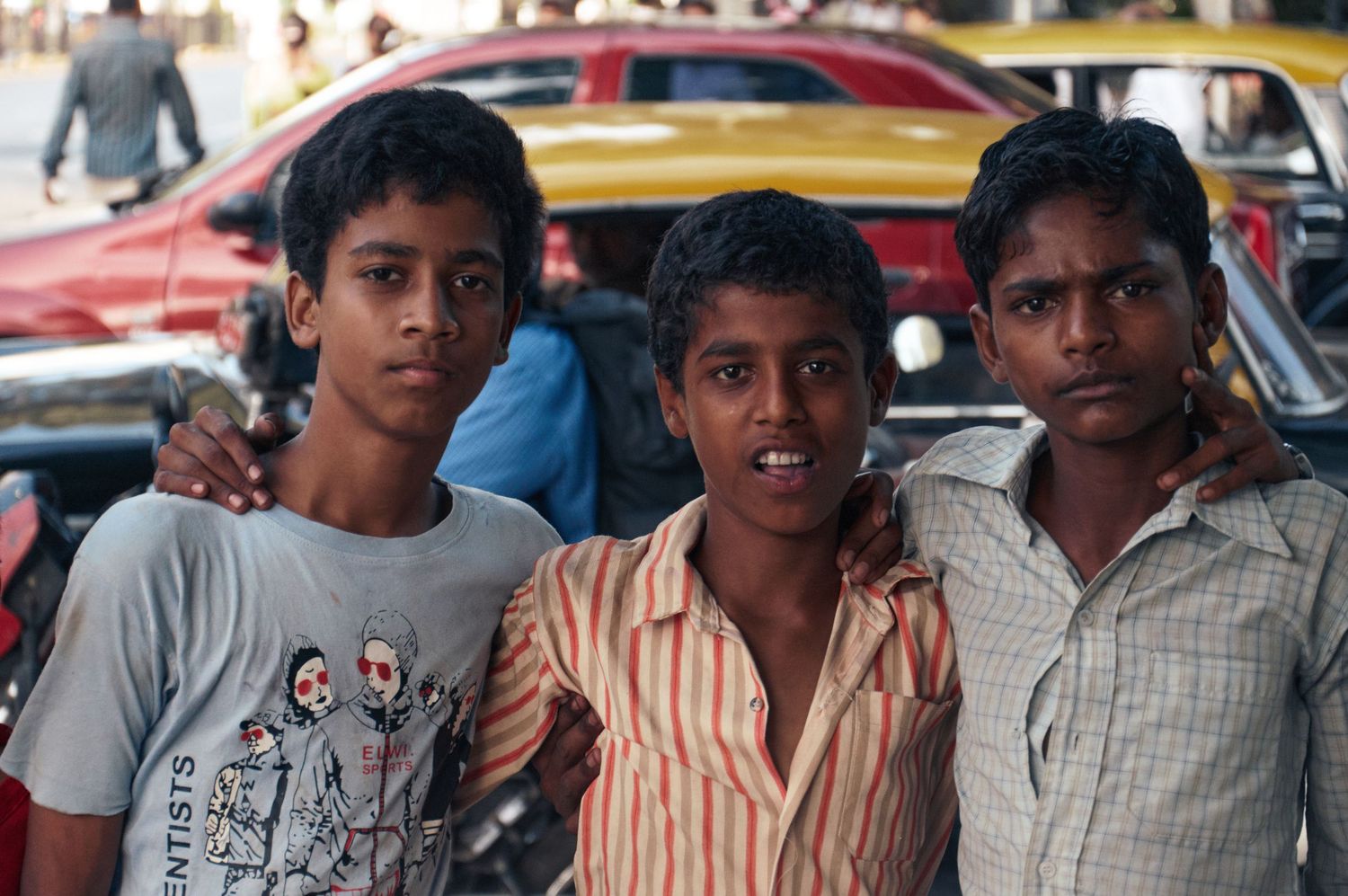 Three young boys posing together on a busy street in India, smiling at the camera with vehicles in the background.