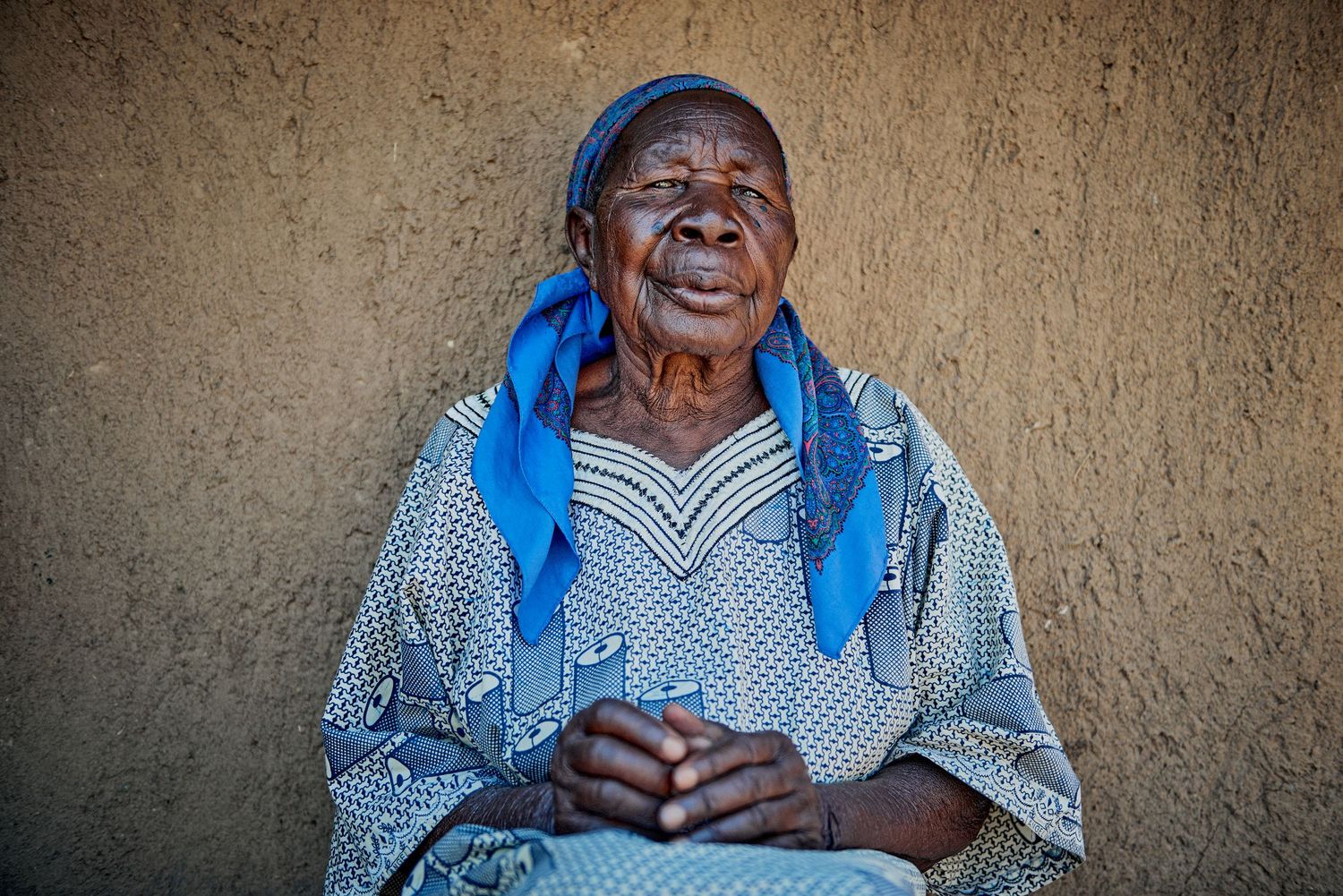 Elderly African woman in blue headscarf and patterned dress sitting against a mud wall, smiling warmly.