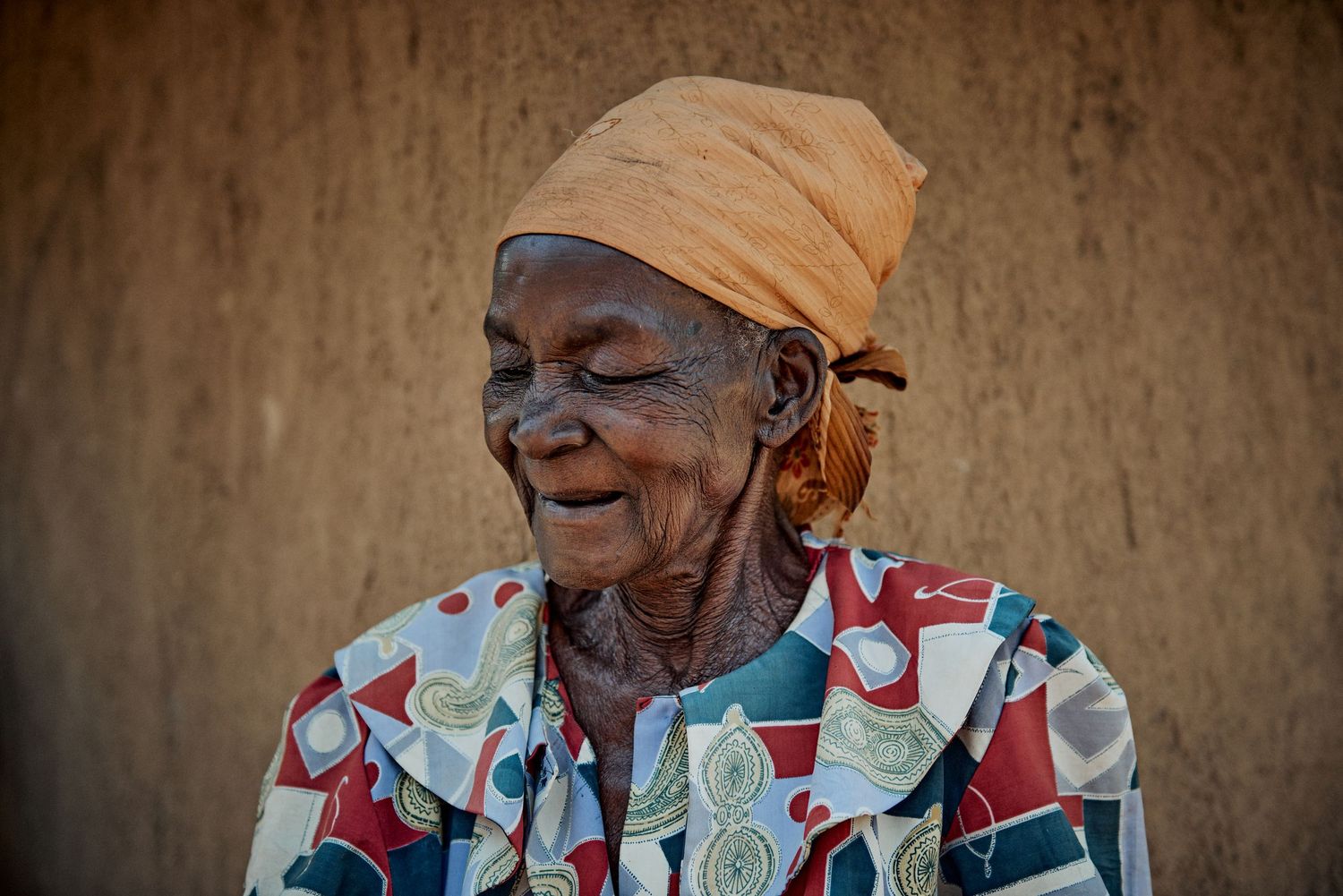 Elderly African woman smiling warmly, wearing colorful floral blouse and orange headscarf, with traditional facial markings.
